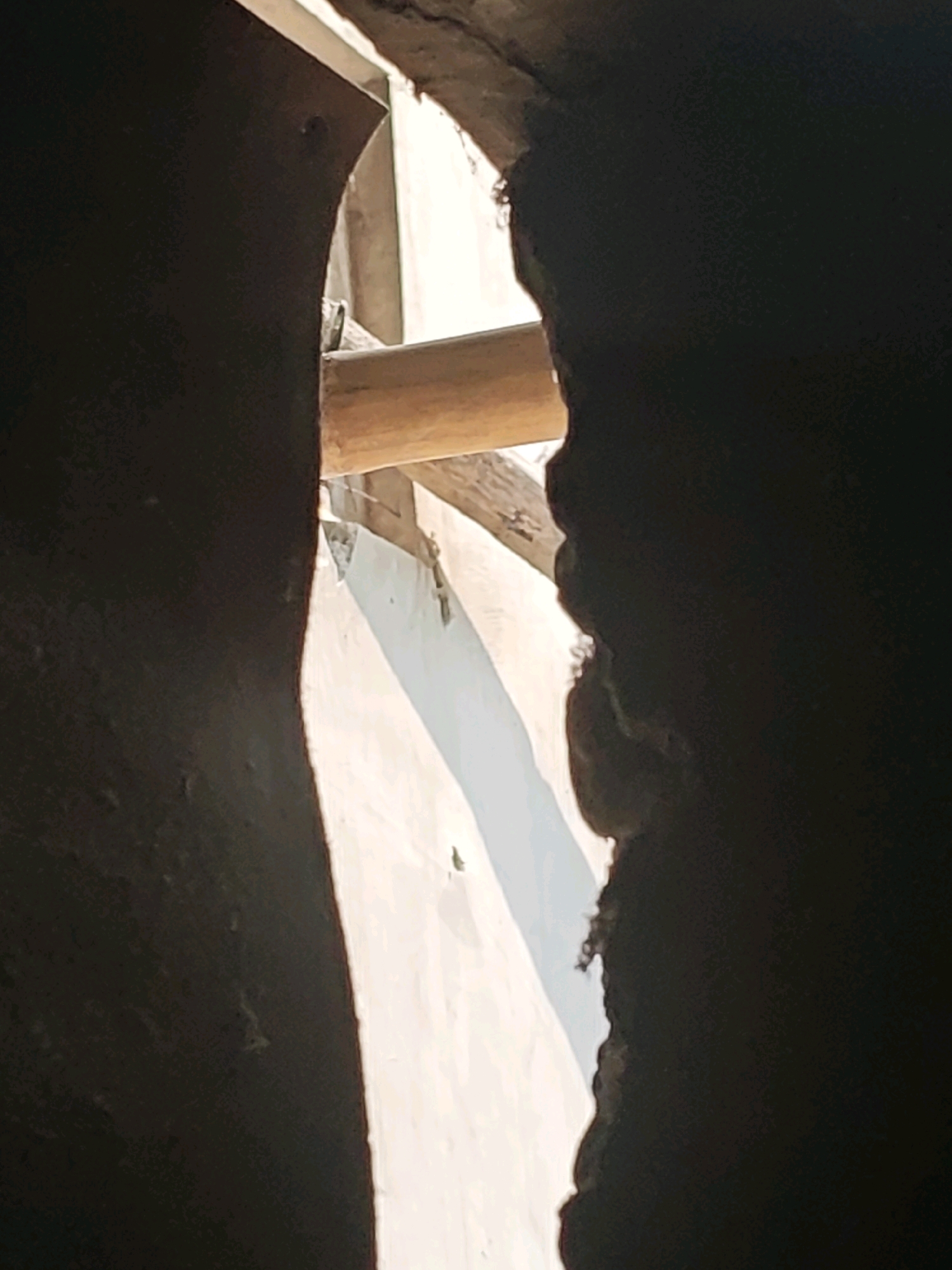 The image shows a narrow crevice between two dark, textured surfaces. A wooden beam is visible across the top of the crevice, casting a shadow. The background is a light-colored wall with a diagonal shadow. There are no people or animals in the image, and no visible signs of specific location within Pagu, Indonesia. The scene appears to be an interior or sheltered outdoor space.