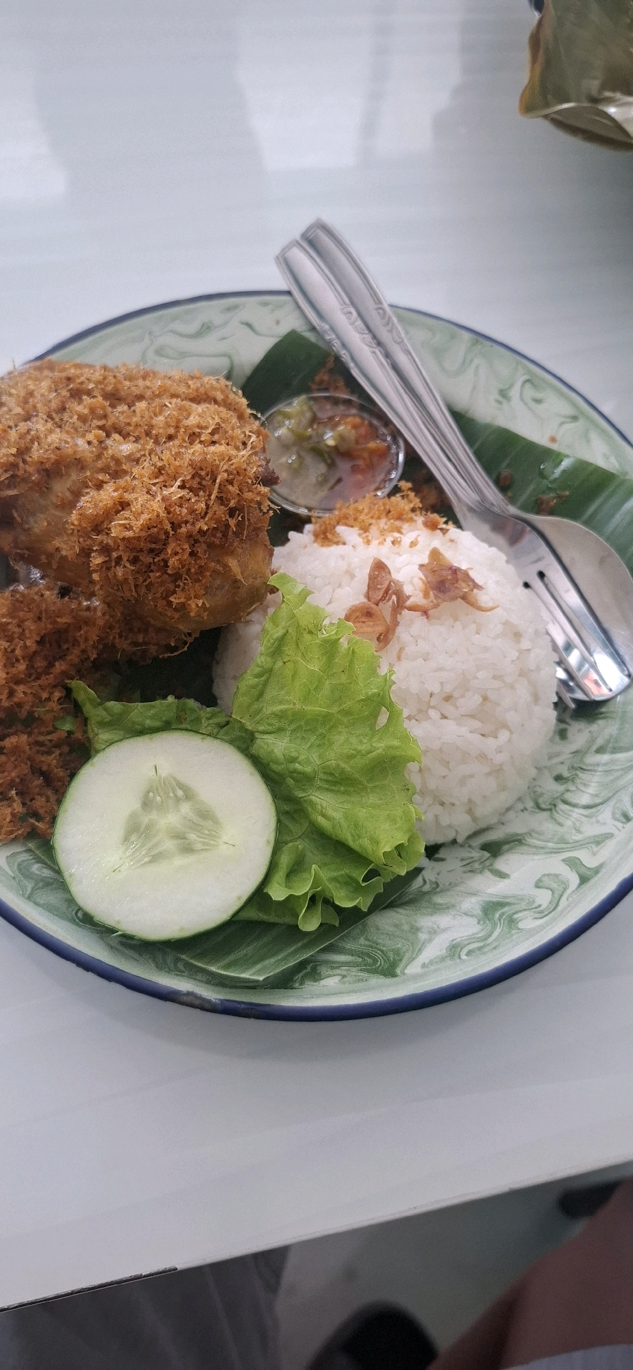 A close-up, high-angle shot shows a plate of Indonesian food, likely from a restaurant in Surakarta, Indonesia. The plate, with a green and white swirled pattern, contains a portion of fried chicken coated in crispy, shredded batter, a mound of white rice, a slice of cucumber, a lettuce leaf, and a small dish of sambal (chili paste). A fork and spoon rest on the plate, partially obscuring the food. The background is blurred, suggesting the photograph was taken indoors in a brightly lit setting. The food appears to be freshly prepared and ready to eat.