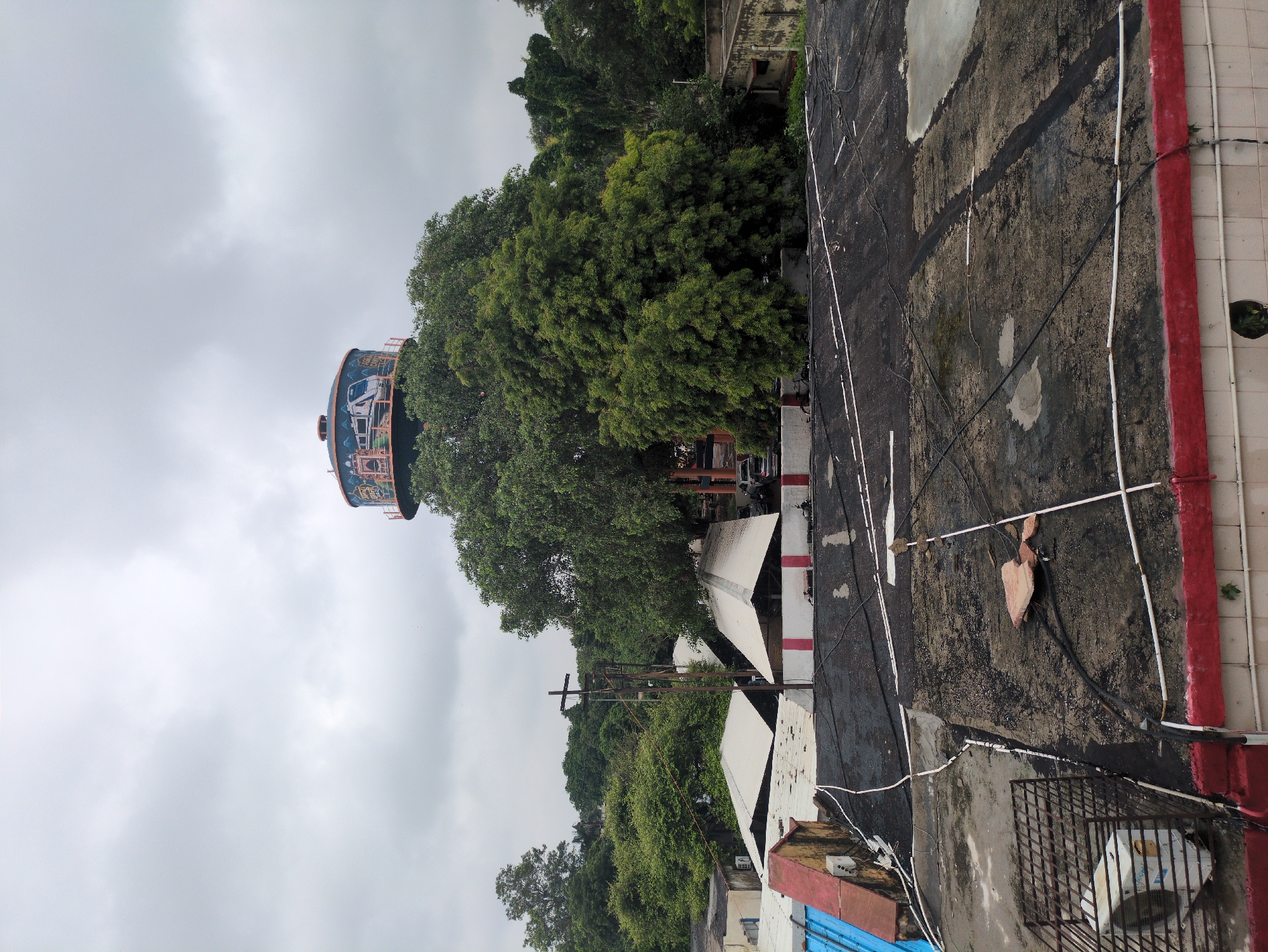 This image captures a scene from Agra, India, featuring a large water tank adorned with a mural of a train, towering over lush green trees. In the foreground, several rooftops are visible, some with a weathered, dark tar-like surface, crisscrossed with white and black electrical wires. One rooftop has a metal cage structure with an air conditioning unit. The buildings below have sloped, white-tiled roofs. The sky is overcast with gray clouds. The overall impression is one of an urban environment integrated with significant natural foliage.