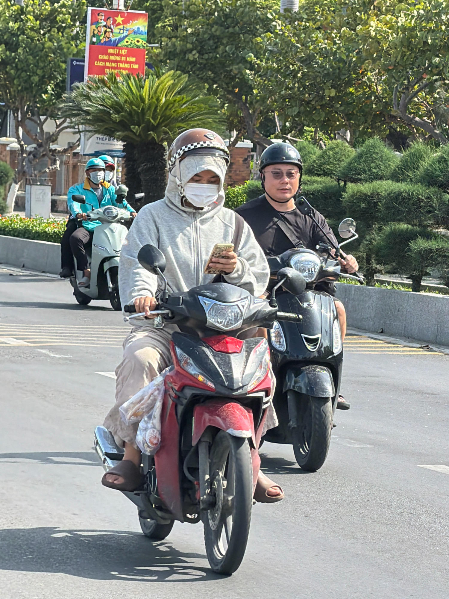 The image depicts a street scene in Nha Trang, Vietnam, with several motorbikes and people. In the foreground, a person wearing a grey hooded jacket, a white mask, and a helmet rides a red motorbike, looking at their phone. Behind them, another person in a black t-shirt and helmet rides a black motorbike. Further in the background, two individuals in teal jackets and helmets are on a light-colored motorbike. A poster with Vietnamese text and images is visible on a pole in the upper left quadrant of the image, alongside trees and greenery. The setting appears to be a sunny day on a paved road with white lane markings.