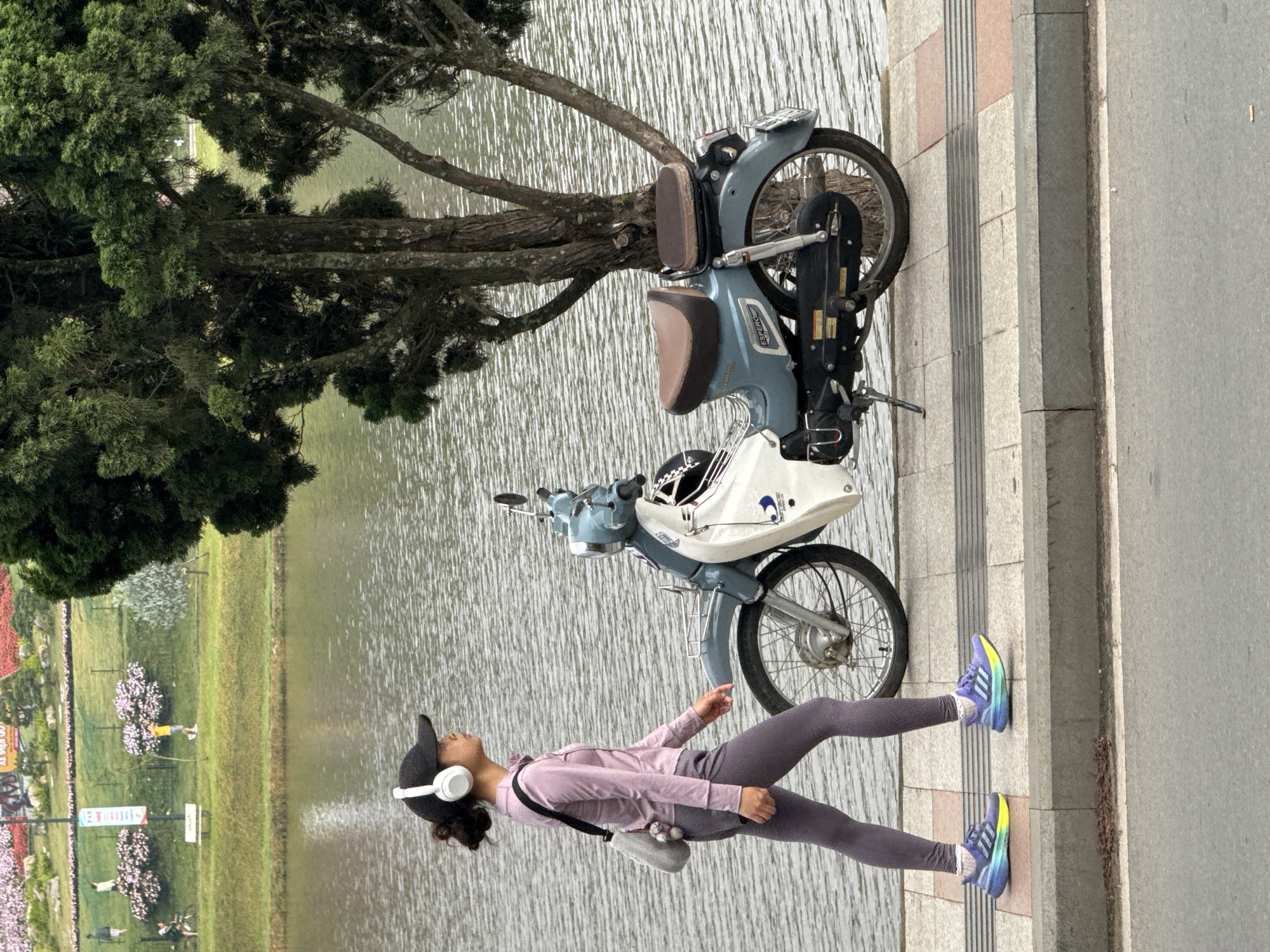 A young woman walks on a sidewalk next to a light blue and white motorbike. She wears a pink long-sleeved shirt, gray leggings, white headphones, and a black baseball cap. Her sneakers are brightly colored with blue, purple, and yellow accents. A grey bag hangs from her shoulder. The motorbike is parked along a textured sidewalk, which runs alongside a body of water that appears to be Xuan Huong Lake in Da Lat, Vietnam, given the surrounding landscape. In the background, lush green grass and trees are visible, along with a colorful flower garden. The overall scene depicts a peaceful, everyday moment in the city of Da Lat.