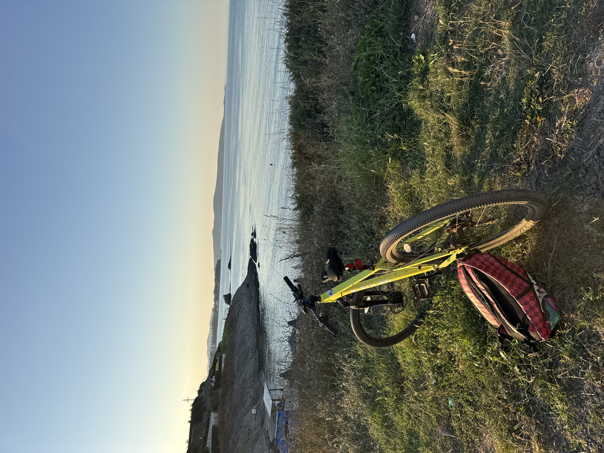 A yellow bicycle with a red and black checkered backpack leaning against it is positioned on a grassy incline overlooking the sea. The bicycle's front wheel is turned to the left. In the background, the sea stretches towards the horizon under a clear blue sky that transitions to a pale yellow near the horizon. Rocky outcrops and a small beach area with some structures and umbrellas are visible along the coastline. Distant hills form the backdrop. The scene is illuminated by the soft light of either sunrise or sunset. The location is identified as Lozenets, Bulgaria.