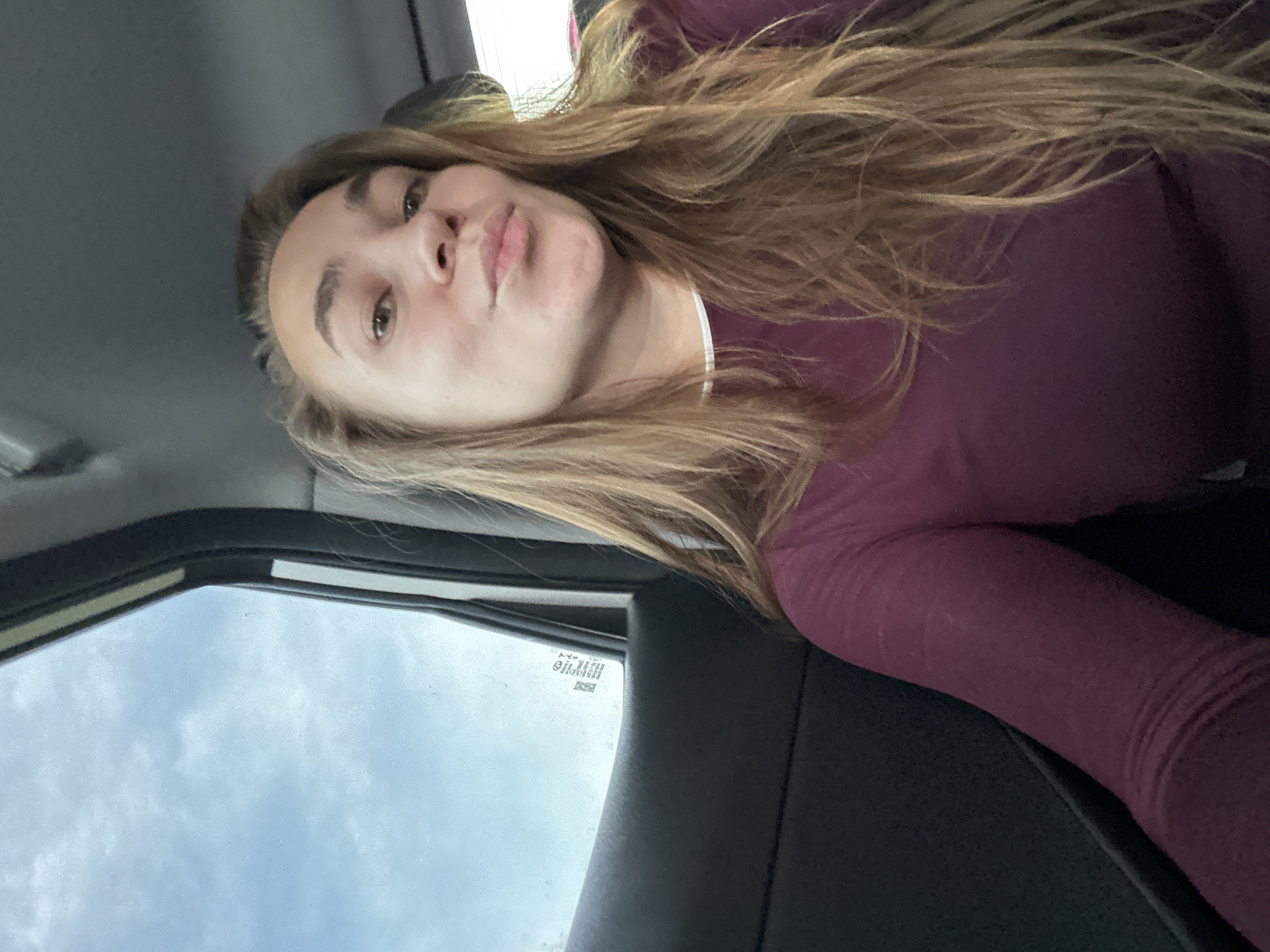 A woman is sitting in the passenger seat of a vehicle, looking at the camera. She has long, blonde hair and is wearing a maroon long-sleeved shirt. The background shows the sky through the car window and the interior of the car. The scene is in Ryabinovka, Russia.