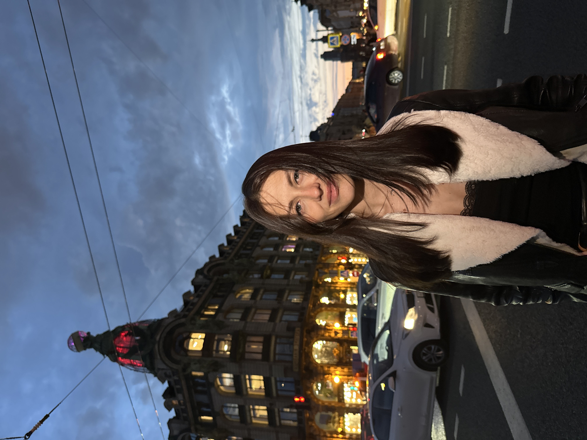 In Saint Petersburg, Russia, a young woman with long, dark hair stands in the foreground, facing the camera. She is wearing a black leather jacket lined with white shearling, and a black lace-trimmed top. Her expression is neutral. Behind her, a street scene unfolds at dusk. Cars are visible on the wet road, with headlights and taillights illuminated. In the background, a grand, ornate building with a domed roof and glowing windows dominates the skyline. The sky is a deep blue, transitioning to hues of orange and purple near the horizon, with scattered clouds. The overall atmosphere suggests an urban evening in a historic European city.