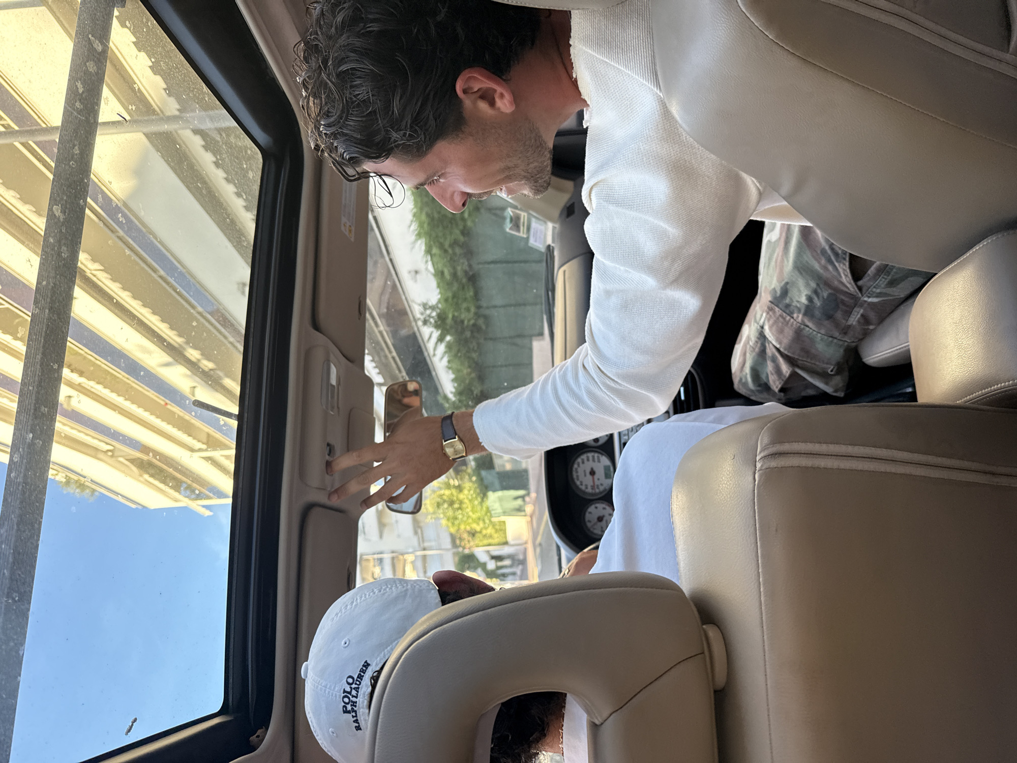 In Cannes, France, two individuals are inside a car with an open sunroof. The person in the foreground, viewed from the back, is wearing a white baseball cap with "POLO RALPH LAUREN" printed on it and a white t-shirt. The driver, on the right, is looking to the right and adjusting something near the sunroof with their right hand. They are wearing a white long-sleeved shirt and camouflage pants. The car's interior is visible, including the dashboard with two circular gauges, and the beige leather seats. Outside the car, buildings and green foliage are visible. The sky is clear and blue. The scene appears to be captured during daylight.