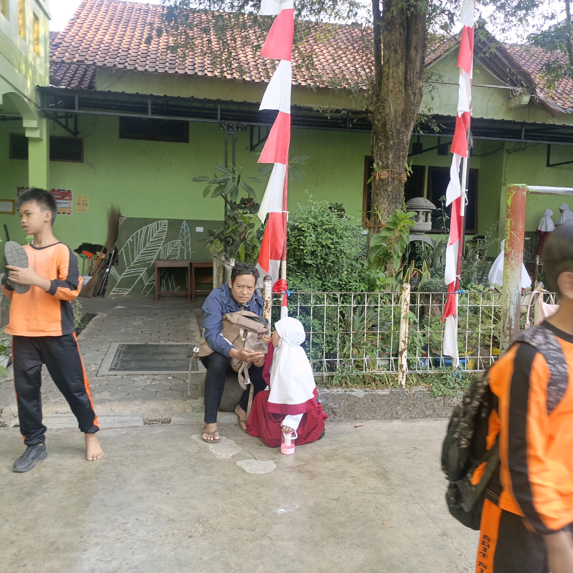 The image depicts an outdoor scene in front of a building with a green facade and a red tile roof, likely a school or community center in Salatiga, Indonesia. A young boy in an orange and black tracksuit stands on the left, holding a shoe. To his right, an adult man wearing a blue jacket is sitting on steps, holding a backpack and a clear container. A young girl in a white hijab and red dress is sitting next to him, holding a pink water bottle. In the background, a flagpole with Indonesian flags (red and white) is visible, along with trees, foliage, and a white metal fence. The ground is paved with concrete.