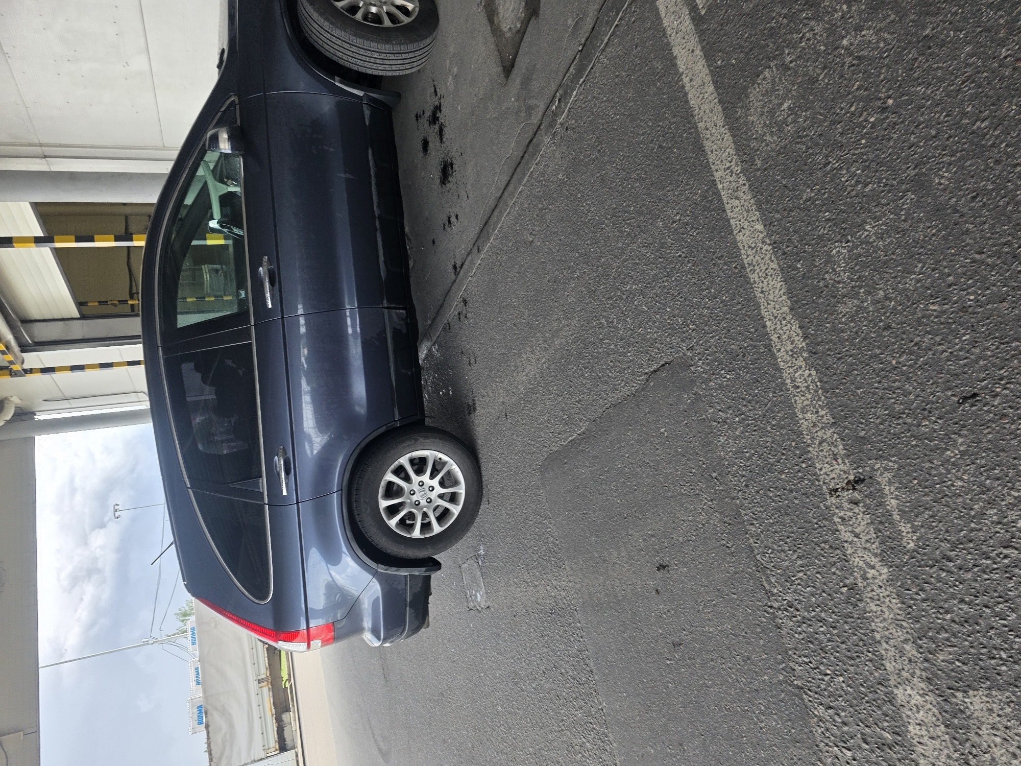 The image shows the side view of a dark blue SUV parked on asphalt pavement. The SUV has tinted windows and alloy wheels. In the background, there is a building with a large garage door. The sky is overcast. To the left of the SUV, a white truck with blue lettering on its side is partially visible. The scene appears to be in an industrial or commercial area.