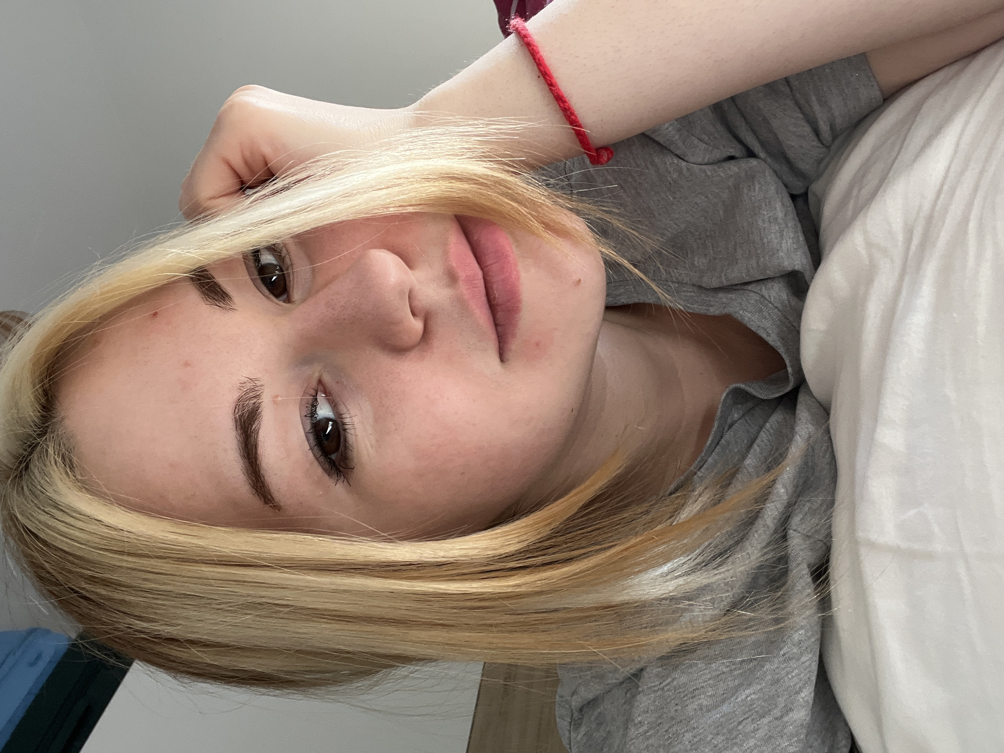 A close-up, angled portrait shows a young, fair-skinned woman with blonde hair lying on a white pillow. She wears a gray t-shirt and a red string bracelet on her wrist. The background includes a white wall. There is no indication that the image was taken in Boreti, Montenegro.