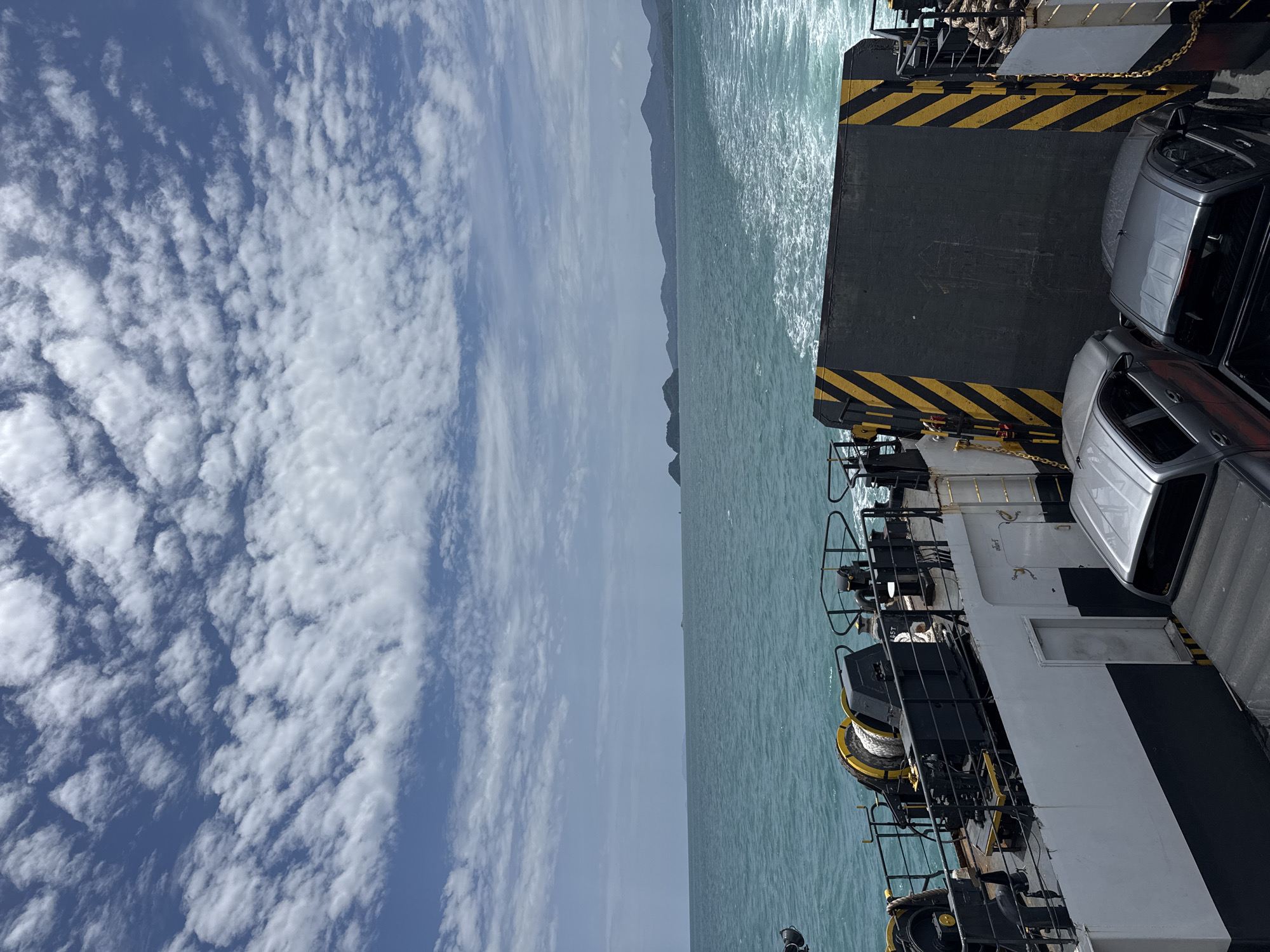 This image depicts a view from the deck of a ferryboat navigating the turquoise waters off the coast of Ko Pha-ngan, Thailand. The sky is a vibrant blue, adorned with scattered white clouds. In the foreground, the deck of the ferry is visible, carrying two pickup trucks. The ferry features prominent yellow and black striped warning signs on its stern. The sea is a calm, wavy expanse, with a visible wake trailing behind the boat. Distant islands with verdant hills dot the horizon. The environment suggests a clear day, ideal for ferry travel between islands. There are no visible people in the image.
