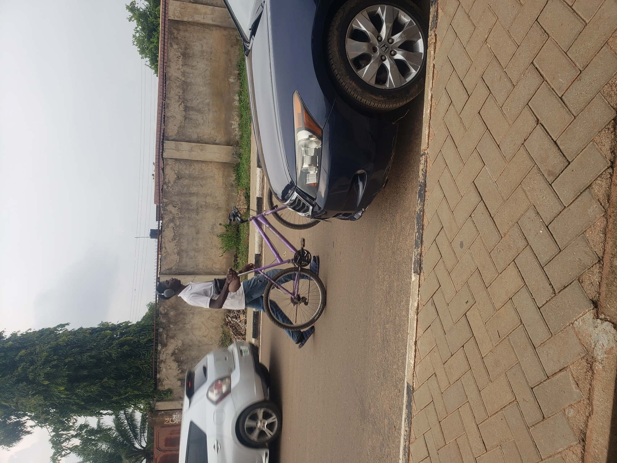 In Narayi, Nigeria, a young Black man wearing a white polo shirt and blue jeans walks across a street, pushing a purple bicycle. He has headphones on and a dark waist pack. The street is paved with asphalt and a brick sidewalk runs along the side. A silver car is passing on the left, and the front of a dark blue sedan is parked on the right. A concrete wall with a textured finish forms a boundary behind the street, with overgrown vegetation visible. The sky is overcast, with a few power lines strung across it.