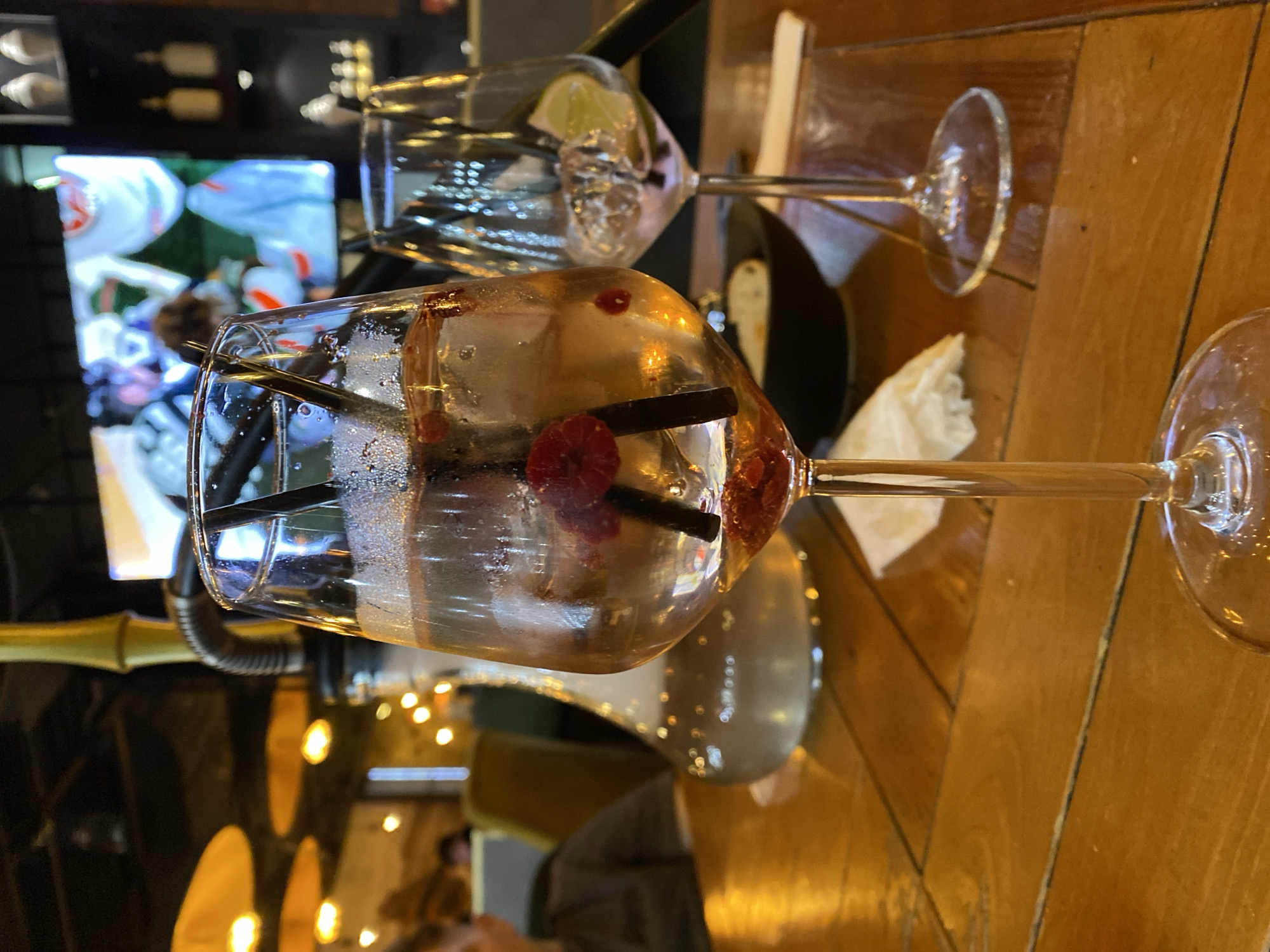 The image shows two wine glasses filled with clear liquid, ice, raspberries, and black straws, positioned on a wooden table. A slice of lime is visible in one glass. In the background, a television screen displays a scene from an American football game. Soft lighting from hanging fixtures is visible, along with partial views of other patrons. The location appears to be inside a bar or restaurant in Yekaterinburg, Russia.