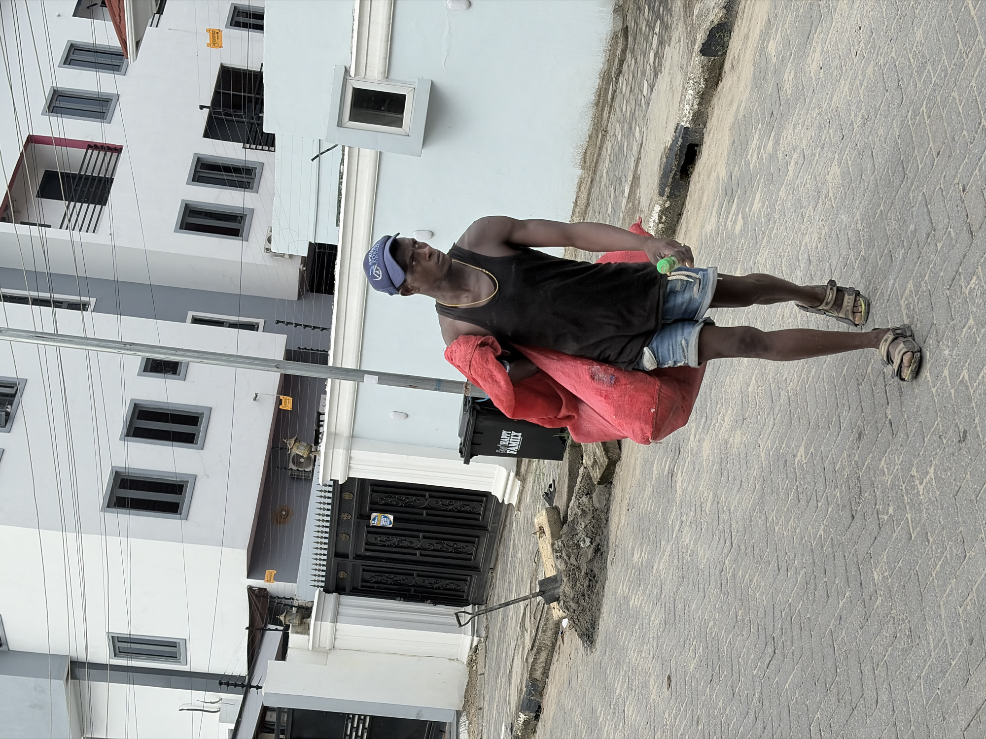 A medium shot depicts a man standing in Igboefon, Nigeria, on a concrete paved walkway next to a multi-story white building. The man is wearing a black tank top, blue shorts, brown sandals, and a blue baseball cap. He is carrying a large red bag over his shoulder and holding a green object in his hand. Next to the man is a black trash can and a shovel. The building behind him has multiple windows and a black gate. Overhead power lines are visible.