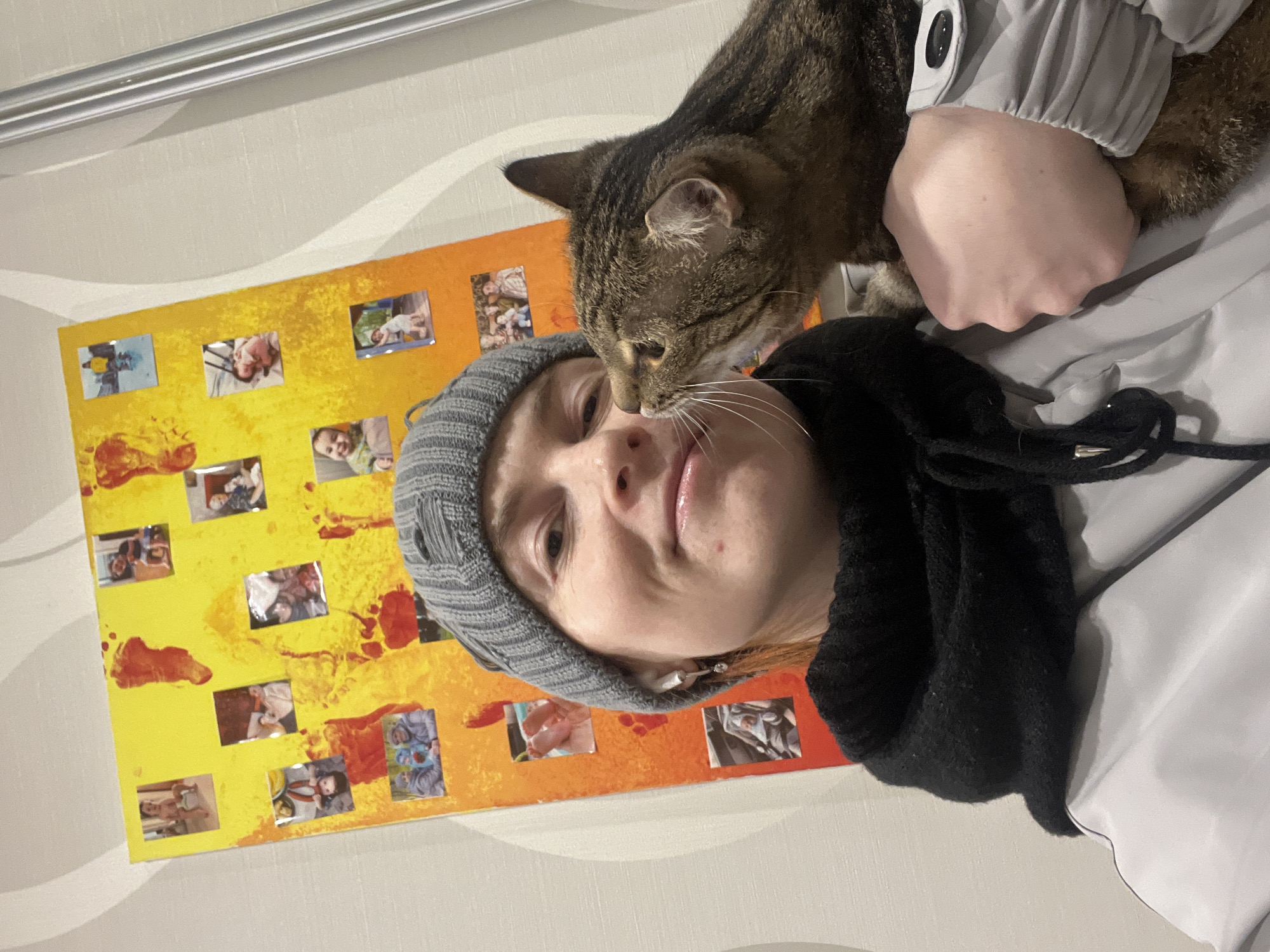 A woman with fair skin and light reddish-brown hair is wearing a grey knit hat and black scarf, holding a tabby cat with brown and black markings. The cat is positioned close to the woman's face, appearing to nuzzle her nose. The woman is wearing a light grey jacket with visible buttons and is holding the cat with her left hand. Behind the woman is a yellow and orange painted board featuring small rectangular photographs adhered to it. The scene appears to be indoors with a light-colored wall in the background. The location is Chaikovsky, Russia.
