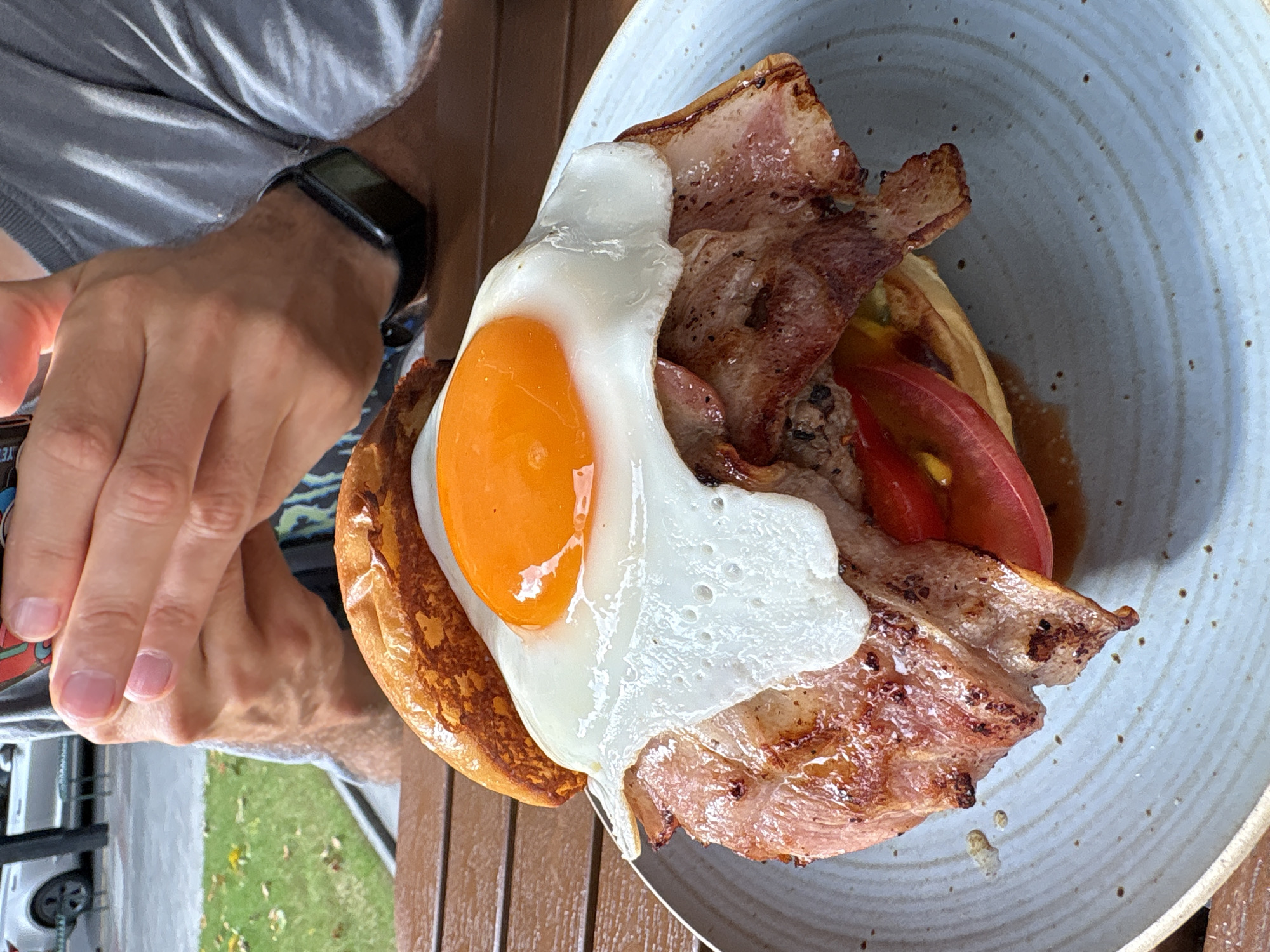 A close-up, eye-level shot shows a burger with bacon and a fried egg on top, resting on a blue-speckled plate. The burger is served on a wooden table, with a blurred background showing a person's hands, a grey t-shirt, and a smartwatch. The person's hands are clasped together, and they appear to be holding something, possibly a phone. The setting appears to be an outdoor cafe or restaurant, with a glimpse of greenery and a parked car in the distance. The overall lighting is natural, suggesting it is daytime. The image does not provide any specific clues to pinpoint its location in Changning District, China.