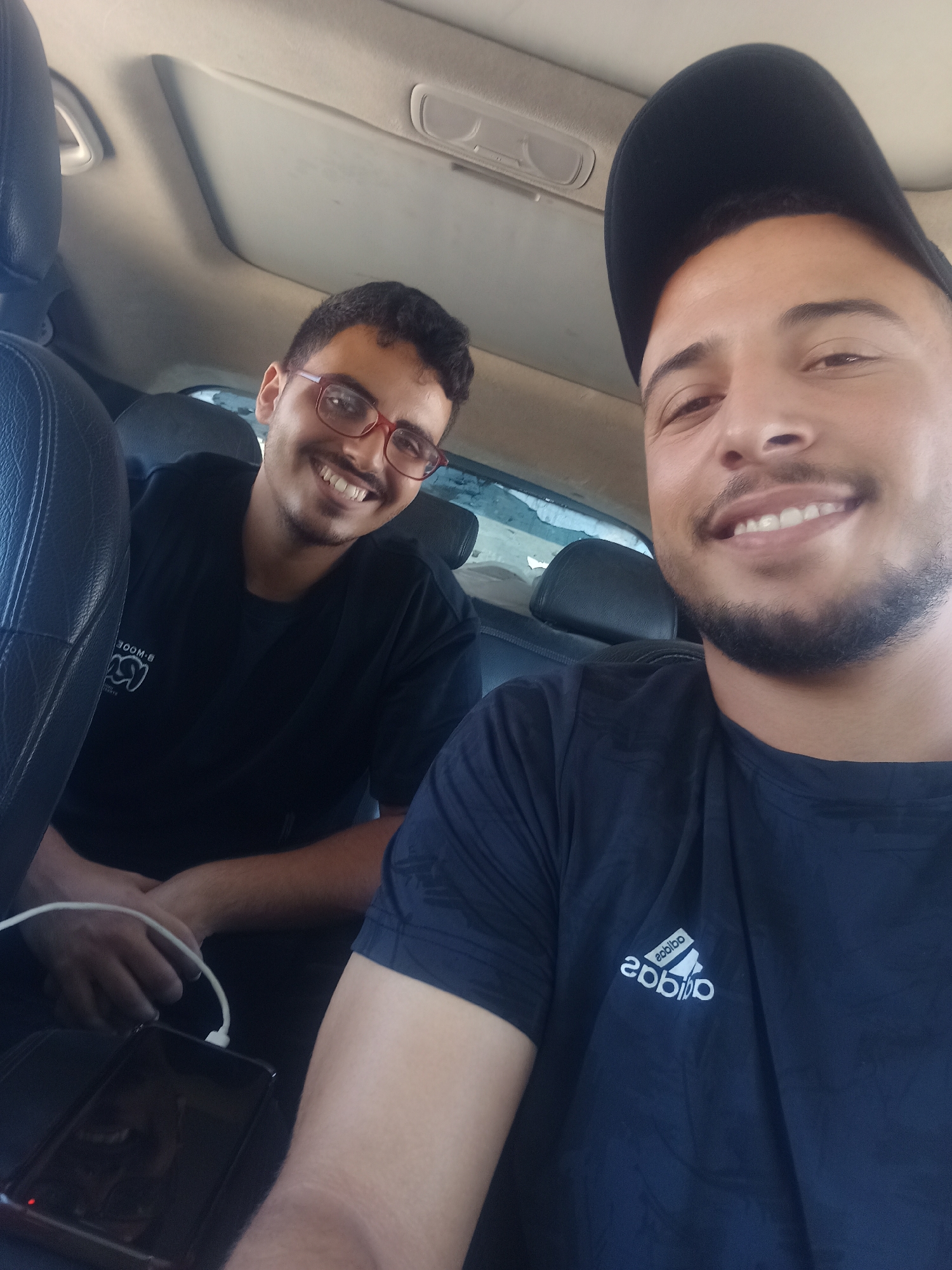 The media file shows two young men inside a vehicle. One man is in the foreground, wearing a black baseball cap and a black t-shirt with an Adidas logo. He is smiling at the camera, with a neatly trimmed beard. The other man is in the background, wearing glasses and a black t-shirt. He is also smiling. A smartphone is visible in the foreground, plugged into a charger. The interior of the car, including the sunroof and seats, is visible, suggesting the photo was taken during the daytime. The overall mood appears cheerful and relaxed.
