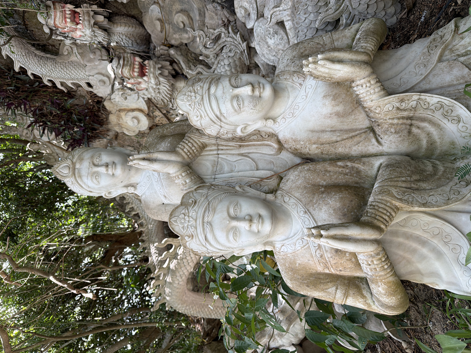 A statue of three figures, likely Buddhist deities, is shown in an outdoor setting. Two figures are in the foreground, kneeling with hands pressed together in prayer. They are adorned with ornate headdresses and flowing robes. Behind them, a taller figure also with hands in prayer stands, looking forward. The statues are made of white stone and show signs of age and weathering. In the background, carved dragons and other mythical creatures are visible, integrated into a rocky, mountainous landscape. Lush green foliage surrounds the statues, suggesting a garden or temple complex. This scene is located at the Marble Mountains, an area of natural and spiritual significance in Đà Nẵng, Vietnam.