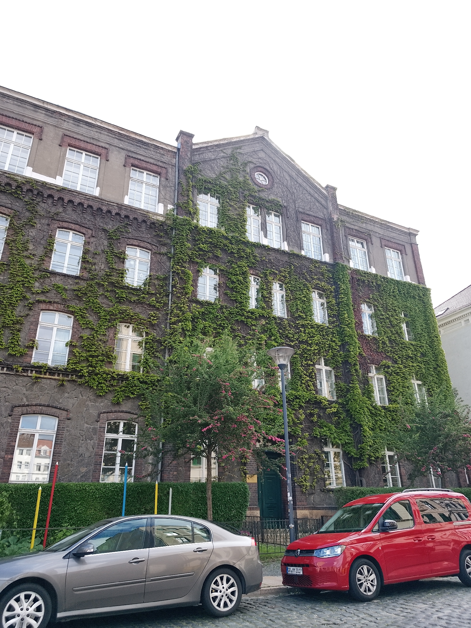 The image captures a four-story building covered in ivy, with multiple windows. The building has a gray brick facade and a distinct architectural style. In front of the building, a silver sedan and a red van are parked on a cobblestone street. A street lamp stands between the two vehicles. A green hedge runs along the front of the building behind the cars, and colorful poles are visible behind the hedge to the left. The location is in the city of Unknown, Unknown.