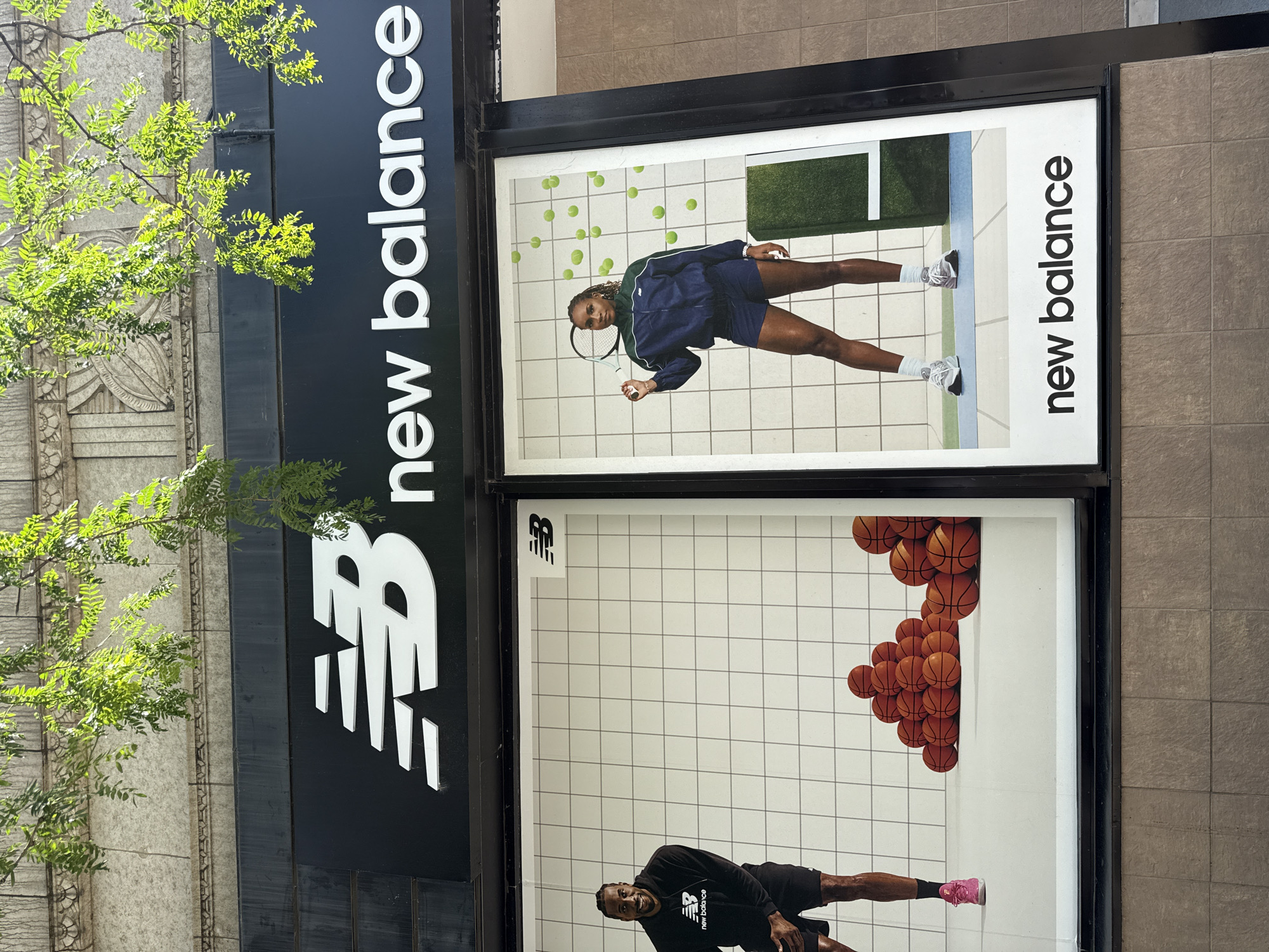 The media file shows the storefront of a New Balance store in Chicago. The store features large window displays showcasing athletic apparel and footwear.
In the upper display, a woman, likely an athlete, is pictured in tennis attire, holding a tennis racket. She is looking at the camera with a neutral expression. Scattered around her are tennis balls, and a blurred green background suggests a tennis court. The "new balance" logo is prominently displayed vertically on the right side of the window.
The lower display features a man, also an athlete, in athletic wear, including a black hoodie with the "New Balance" logo. He is in a dynamic pose, possibly stretching or preparing for activity, with a slight smile. A cluster of basketballs is stacked in the background, and a pair of pink athletic shoes is visible on his foot. The "new balance" logo is again visible vertically on the right.
Above the window displays, the store's name, "new balance," is emblazoned in large white lettering against a black background. A stylized "NB" logo is also visible on the black facade.
The scene appears to be outdoors during the daytime, with bright sunlight casting shadows. Lush green foliage from a nearby tree partially obscures the left side of the store's facade, adding a natural element to the urban setting. The building's architecture suggests an established urban environment.
