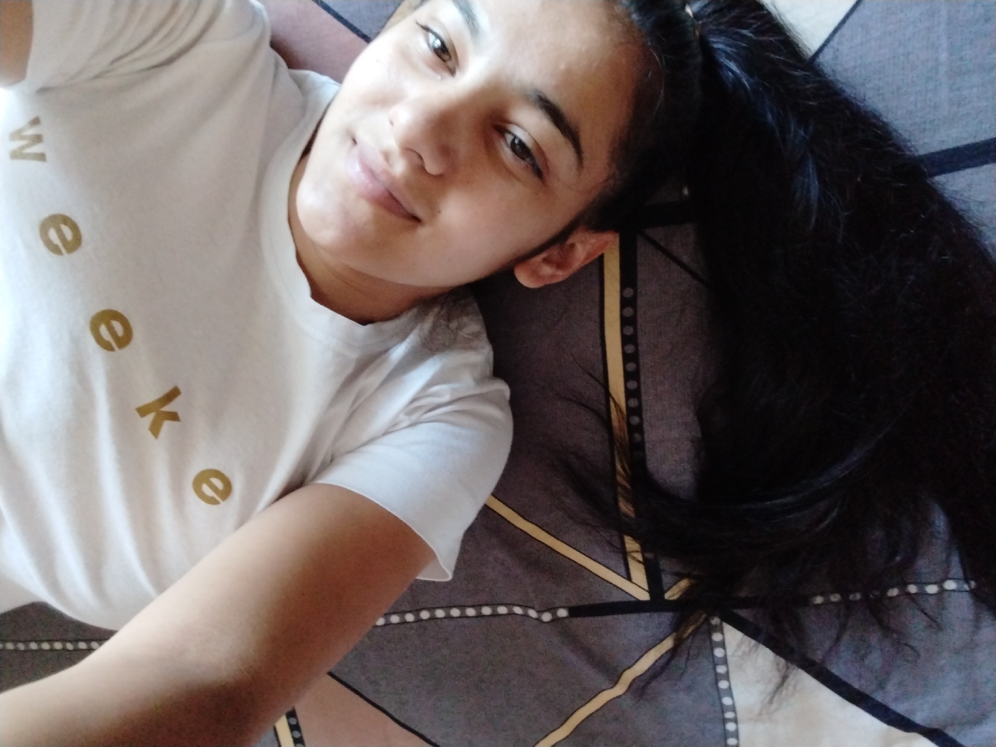 A young woman with long, dark hair is lying on a bed. She is wearing a white t-shirt with the letters "Week" printed on it. The bed has a patterned comforter with geometric shapes and lines. The location is an unknown city and unknown location.