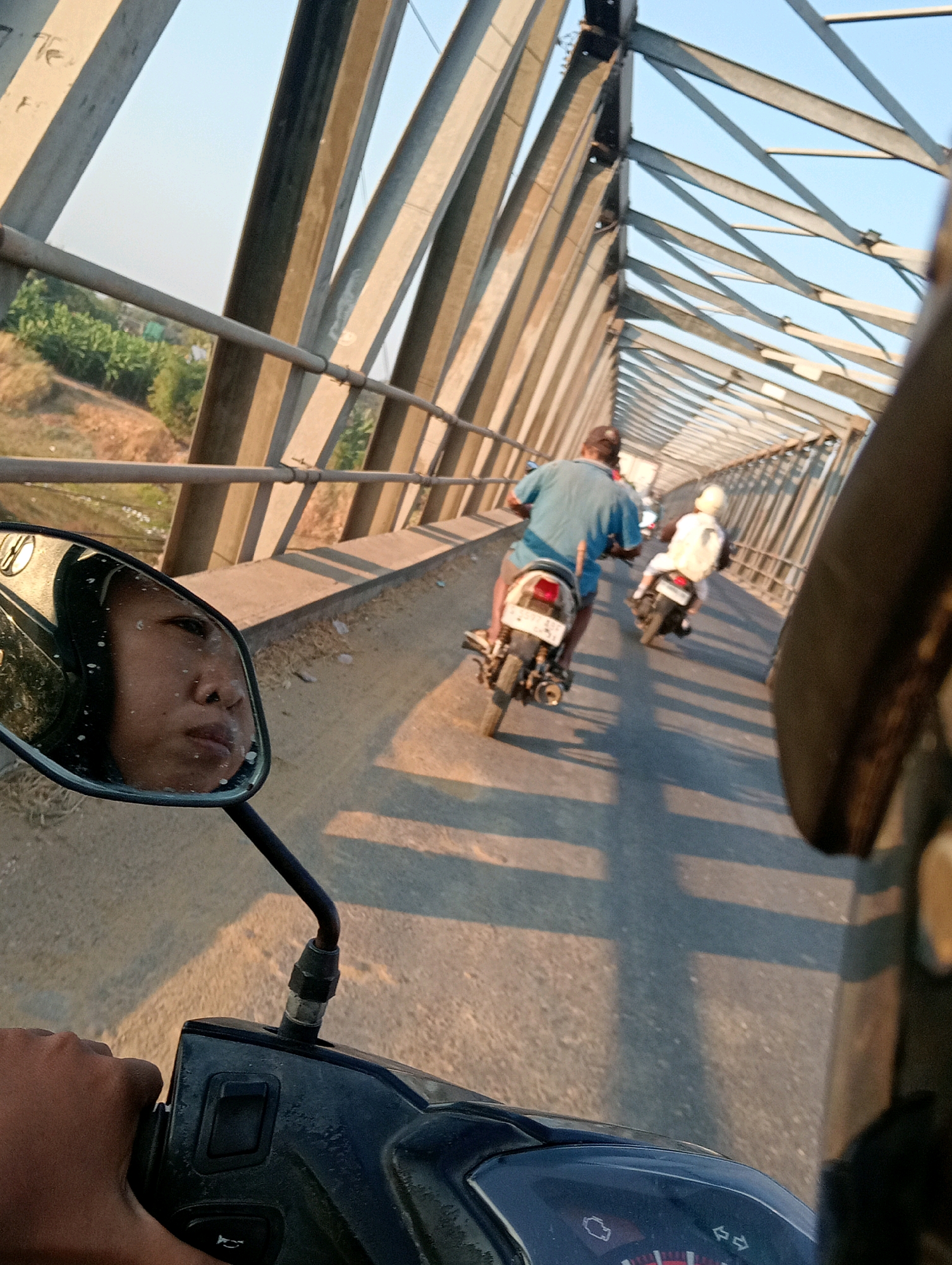 In Soko, Indonesia, two individuals are riding motorbikes across a metal truss bridge. The perspective is from a motorbike driver looking forward and to the left, with their hand on the handlebars. In the mirror on the left, the face of the rider is visible. A second motorbike, carrying a passenger wearing a white helmet and jacket, is ahead and to the right. The bridge spans over a dry, dusty embankment with sparse vegetation. The sky is clear and bright blue, with the sun casting long shadows across the bridge. The metallic structure of the bridge is prominent, with diagonal beams forming a latticework overhead. The road surface is asphalt.