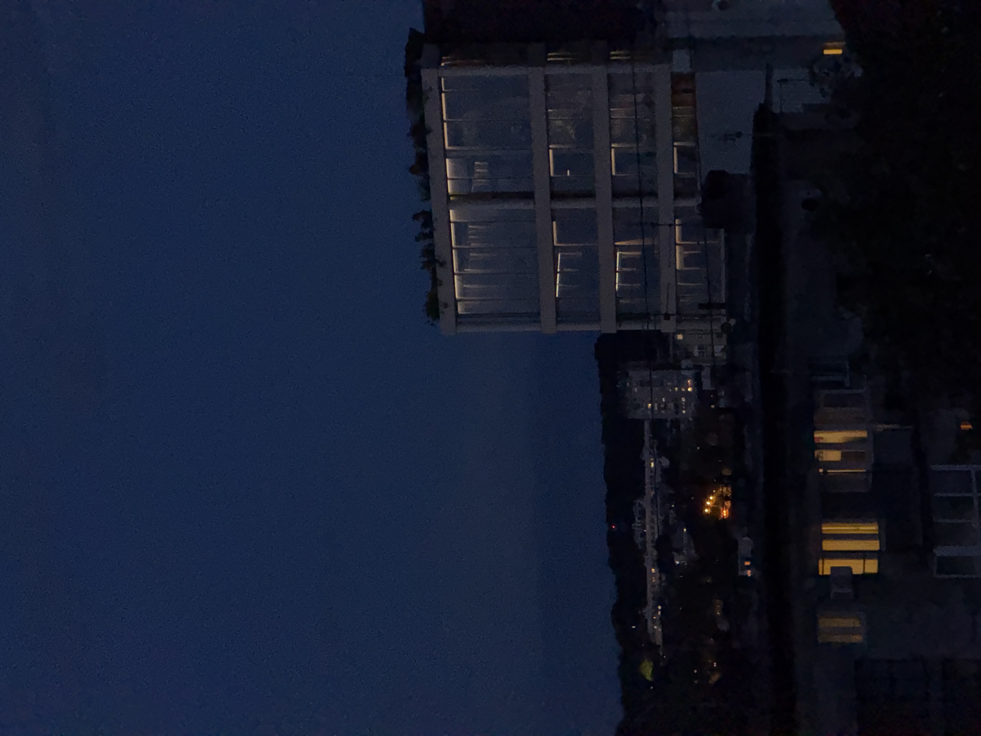 Night view in Vracar Urban Municipality, Serbia. High-rise apartment buildings with illuminated windows visible. Dark sky above. Other buildings and sparse lighting on the street below. Dense vegetation bordering the buildings.