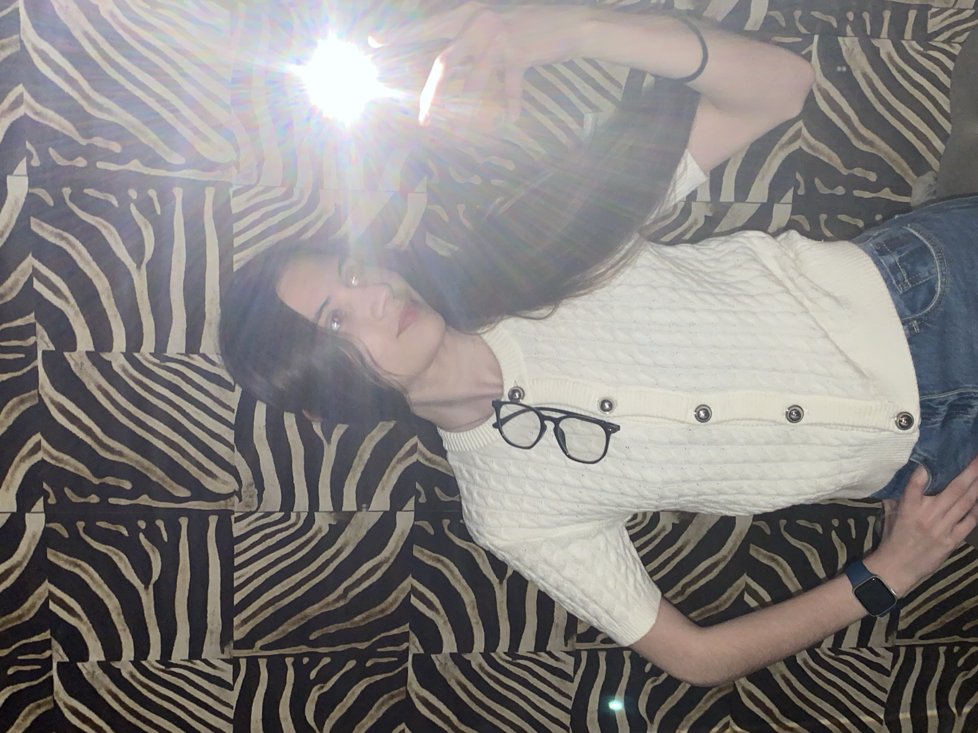An overhead photograph captures a young individual lying on their back on a distinctive black and beige zebra-print surface, which appears to be a patterned blanket or rug. The person has long, dark hair fanned out around their head and is looking directly towards the camera. They are wearing a cream-colored, cable-knit cardigan with four prominent silver, metallic buttons down the front, paired with blue denim jeans. A pair of black-framed eyeglasses is neatly tucked into the neckline of the cardigan. On their left wrist, a dark blue smartwatch is visible.
A very bright, star-shaped light flare, likely from a camera flash, emanates from the upper left portion of the image, partially obscuring the background and creating a strong lens flare effect across the scene. The flash illuminates the person's face, causing a red-eye effect in their pupils. Their right arm is extended upwards, suggesting they may be holding the device that captured the image. The person's expression is neutral and calm. There is no visible text in the image. The scene suggests an indoor setting, and the provided context indicates the location is Votkinsk, Russia.