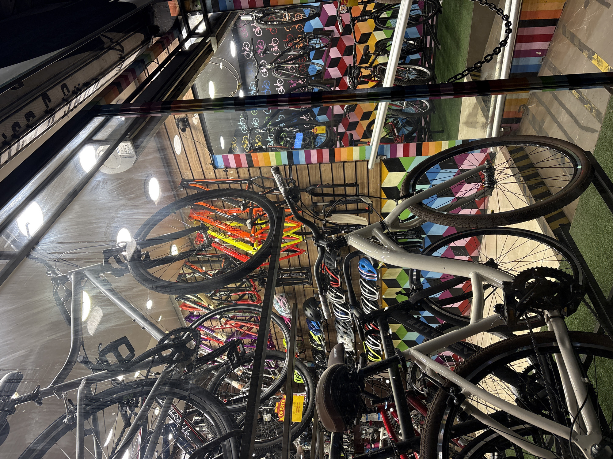 The image depicts the interior of a bicycle shop, likely located in the city of São Paulo, Brazil, as suggested by the context provided. The shop is filled with various bicycles, some hanging from the ceiling and others displayed on racks. The bicycles are of different colors and models, including what appear to be road bikes, mountain bikes, and possibly city bikes.
In the foreground, a white bicycle is prominently displayed, showcasing its frame, wheels, and drivetrain. To its left, a portion of a red bicycle is visible. Further back, rows of bicycle helmets are arranged on shelves along the wall, in an assortment of colors including black, blue, yellow, and pink.
The background features a wall decorated with a colorful, abstract pattern of stripes and geometric shapes. The shop appears to have a glass storefront, through which the interior is visible, along with reflections of the outside environment. The lighting inside the shop is bright, with visible light fixtures on the ceiling. There are no people visible in the image. The overall impression is that of a well-stocked bicycle retail store.