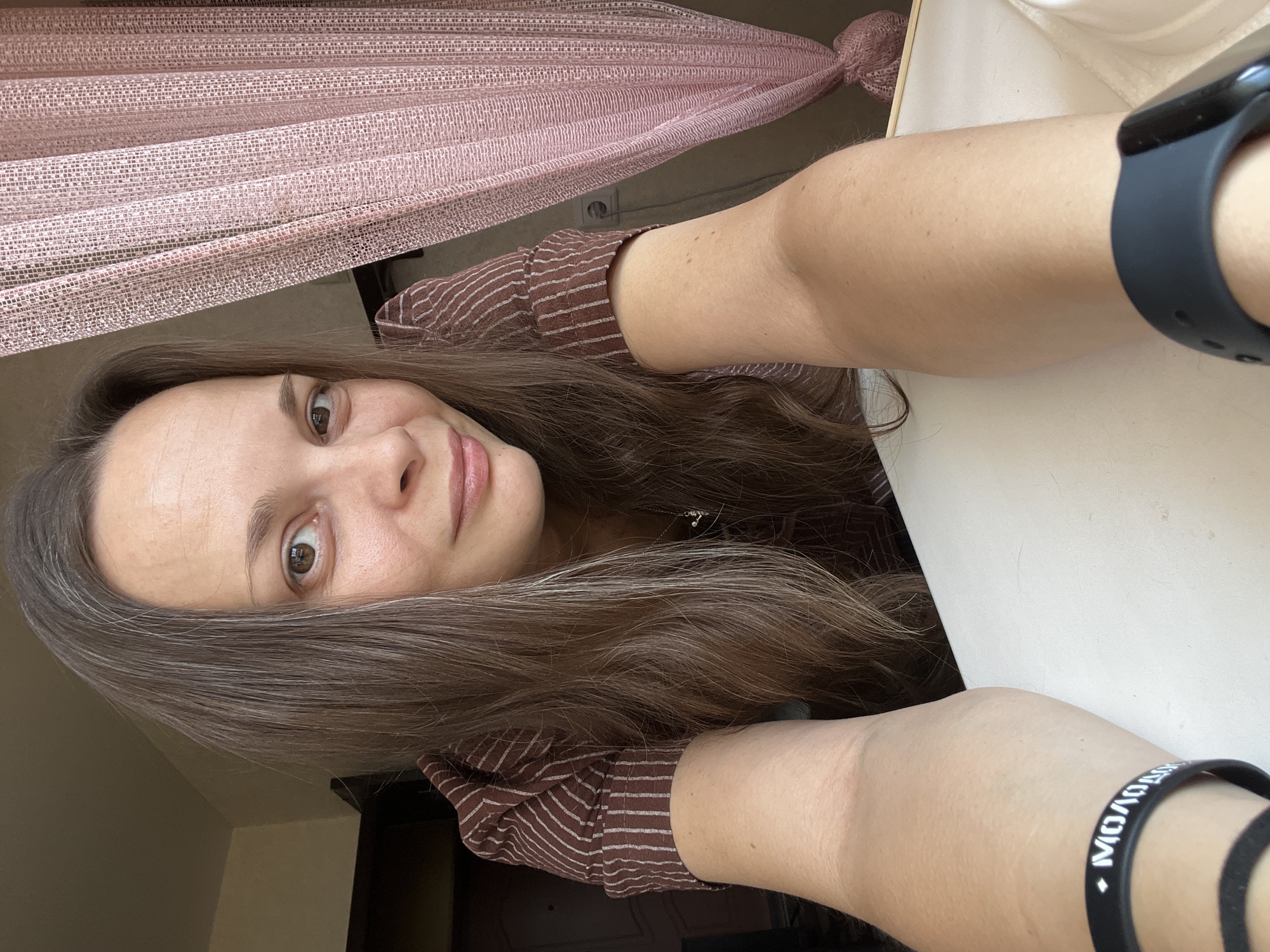A woman with long, brown hair and a light complexion, wearing a brown and white striped shirt, poses for a selfie from a low angle. Her right arm is resting on a white surface, and a black wristband with white Cyrillic text is visible on her left wrist. A pink sheer curtain hangs to the right of the frame, and a portion of a wall with an electrical outlet is visible behind it. The background suggests an indoor setting, possibly a room in an apartment in Tolyatti, Russia.