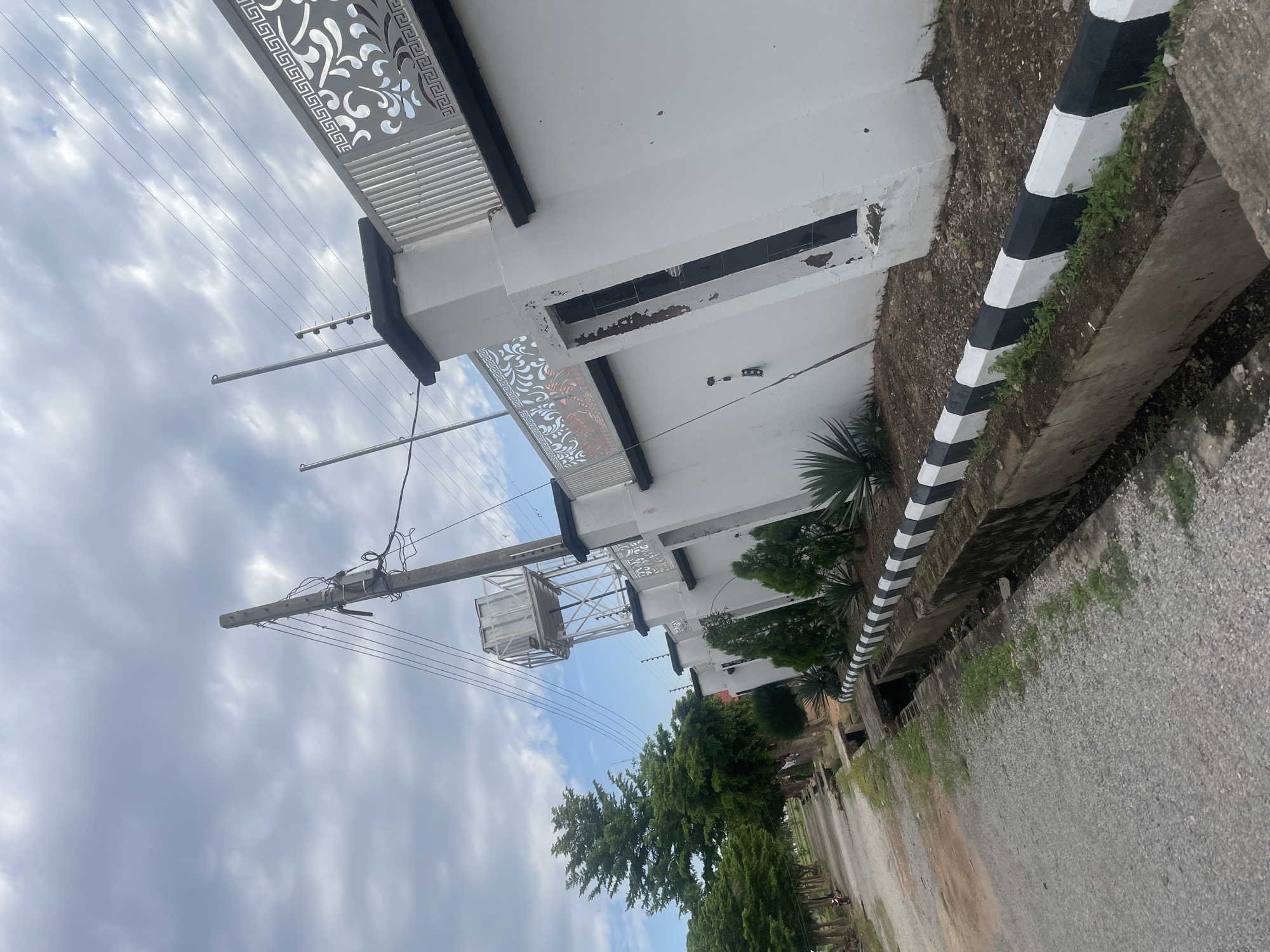 The image depicts a street scene in Jauro Yinu, Nigeria. On the right side of the frame is a white wall with decorative metal railings on top, featuring a black trim. The wall is punctuated by pillars, one of which has a visible opening that appears to be a doorway or window, with black tiles inside. A water tank is elevated on a metal structure behind the wall, supported by a wooden utility pole with electrical wires. The wall is bordered by a drain with concrete edges painted with alternating black and white stripes. In front of the wall, there are some sparse green plants.
The left side of the image shows a paved road or path, with a line of trees running alongside it. Further down the road, there are glimpses of other trees and vegetation. The sky is partly cloudy with a mix of blue sky and grey clouds. A single red motorcycle is parked on the left side of the road, near the trees. The overall impression is that of a semi-urban or suburban residential area.