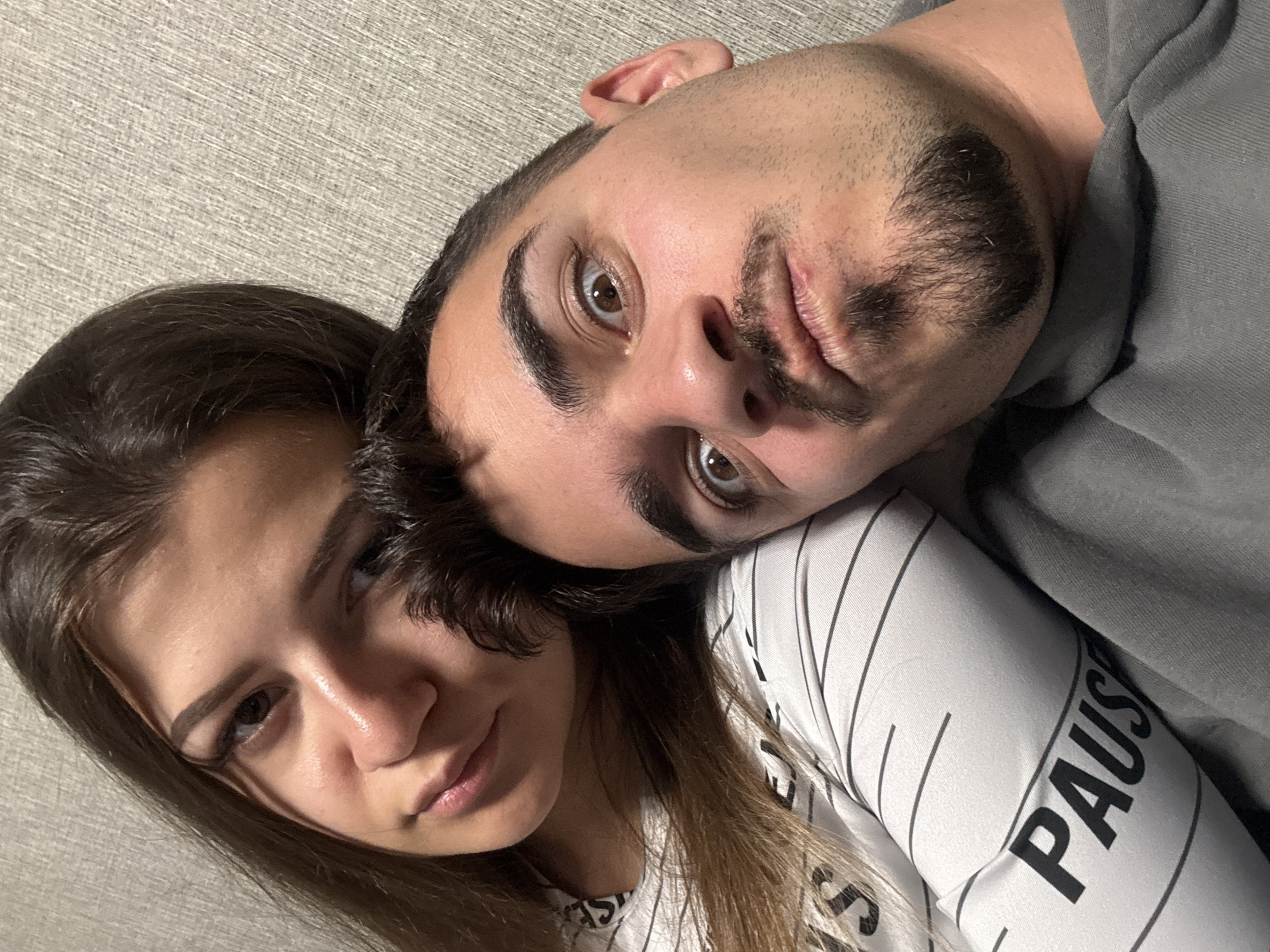 This close-up image captures two young adults, a male and a female, in an intimate indoor setting. The scene is likely a selfie or a portrait taken from an overhead perspective, with both subjects looking towards the camera while lying down or leaning back against a textured, light grey or off-white background, possibly a wall or headboard. The context of the location is Sofia, Bulgaria.
The male, positioned higher and to the right, has short dark hair, a beard, and a mustache. His brown eyes are wide and focused directly on the camera, and he has a neutral expression. He is wearing a plain grey sweatshirt.
The female is positioned lower and to the left, with her head resting on the male's shoulder or chest, indicating closeness. She has long, dark hair and a softer, more relaxed expression, with her gaze directed slightly off-camera. She is wearing a white long-sleeved shirt with thin black vertical stripes. Clearly visible on her sleeve is the word "PAUSE" in bold black capital letters, with an additional "S" seen below it.
The lighting appears to be artificial, casting gentle shadows that define their facial contours. The overall mood of the image is calm and intimate, focusing on the connection between the two individuals. There are no specific cues for time of day or weather, but the indoor setting suggests a controlled environment.