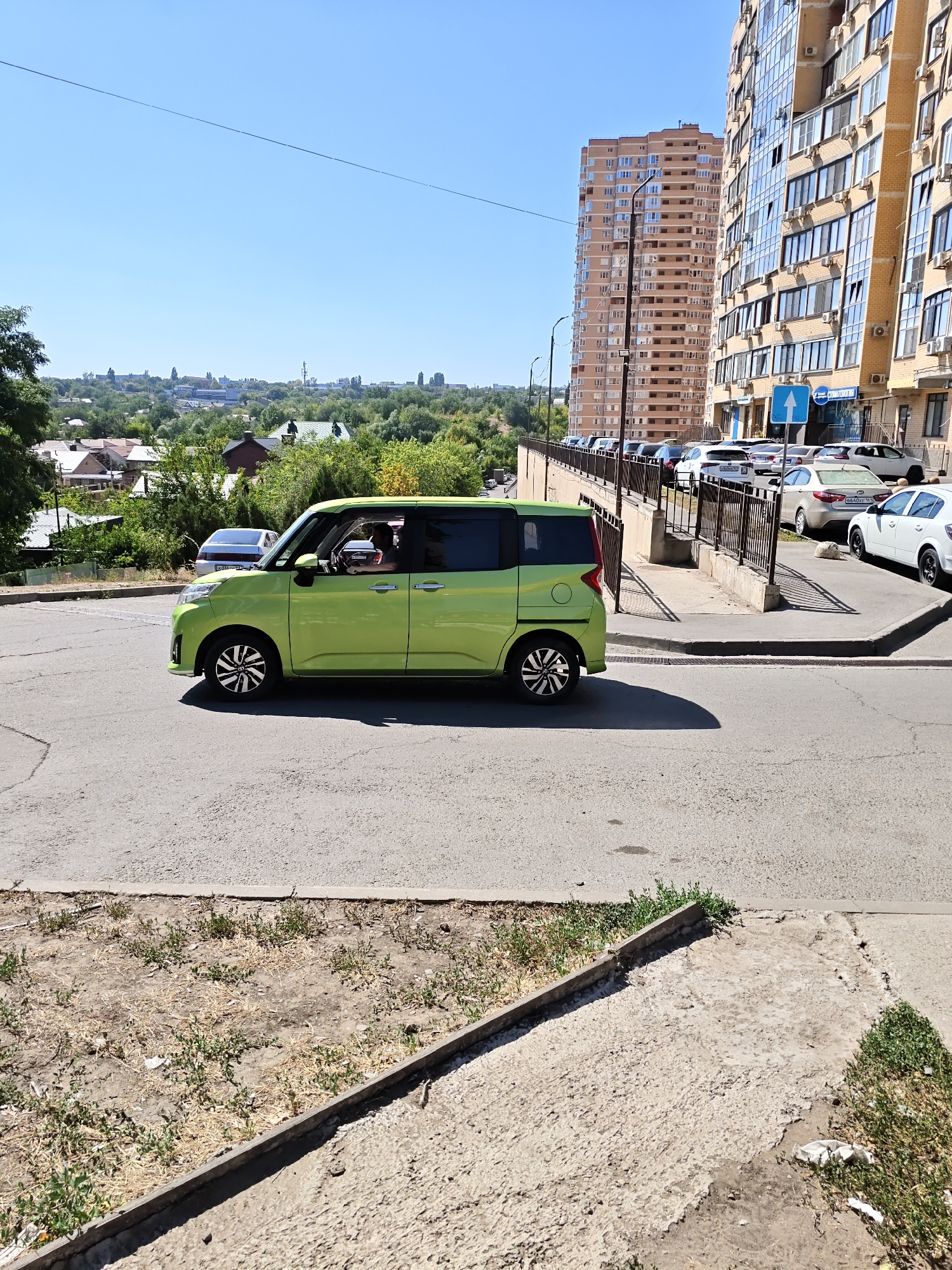 The image depicts a street scene in Rostov-on-Don, Russia, on a clear, sunny day. In the foreground, a bright green hatchback car is moving along an asphalt road. The car has tinted windows, and a person is visible inside, driving. The road has visible cracks and some sparse vegetation growing along its edge.
Behind the car, a raised sidewalk runs parallel to the street, bordered by a metal fence. A row of parked cars is visible on the other side of the sidewalk, beyond the fence. In the background, a dense cluster of trees and houses with red roofs can be seen on a sloping hill. Further in the distance, two tall, modern apartment buildings dominate the skyline, characterized by their beige and light brown facades, numerous windows, and balconies. The sky is clear blue. The scene is consistent with an urban environment with residential areas and modern infrastructure.