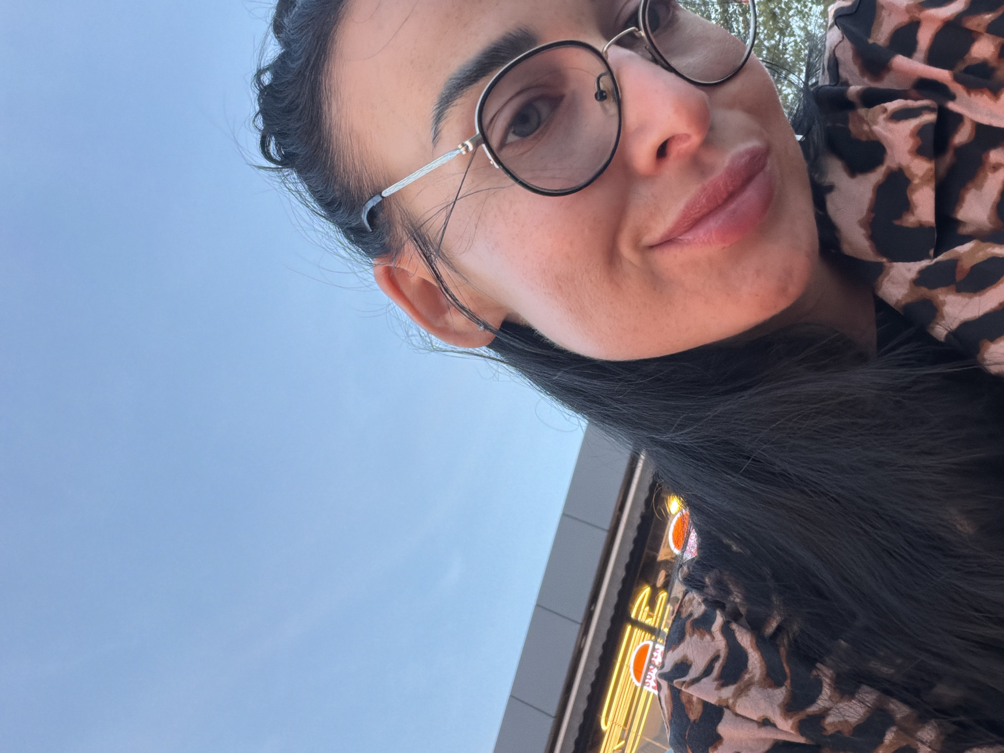 A woman is taking a selfie outdoors, with the sky and part of a building visible in the background. She is wearing glasses and a leopard-print shirt. The sky is a clear blue, and the building appears to be a commercial establishment with neon lights. The overall impression is a casual, outdoor setting. It is not possible to determine the specific location within Sochi, Russia from the provided image.