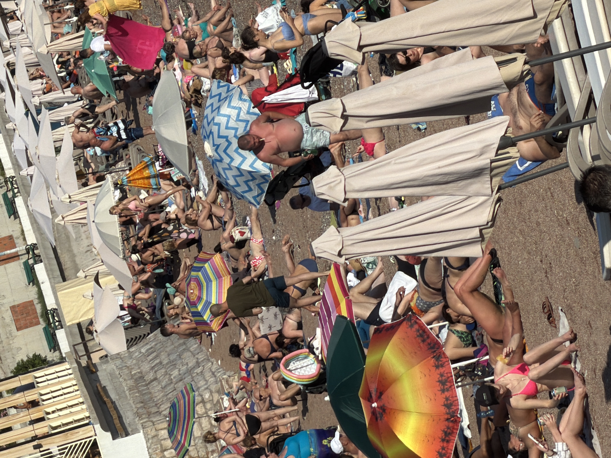 The image depicts a crowded beach scene in Petrovac centar, Montenegro, likely during peak tourist season given the density of beachgoers and umbrellas. The ground is covered in pebbles and sand, typical of a Mediterranean coastline. Numerous beach umbrellas, some brightly colored and others plain beige, are spread across the scene, providing shade to people lounging on beach chairs and towels.
A variety of individuals are visible, mostly dressed in swimwear, with many sunbathing, relaxing, or engaged in conversations. The age range appears diverse, with both adults and some younger individuals present. Some people are partially obscured by umbrellas or other beachgoers.
Objects scattered around include beach chairs, towels, bags, and drinks. A few individuals are captured in motion, such as walking or standing. The backdrop includes a stone wall, possibly part of the town's infrastructure, and some buildings, suggesting an urban setting adjacent to the beach. The lighting indicates a sunny day, with shadows cast by the umbrellas and people. The overall atmosphere is one of leisure and recreation on a busy beach.