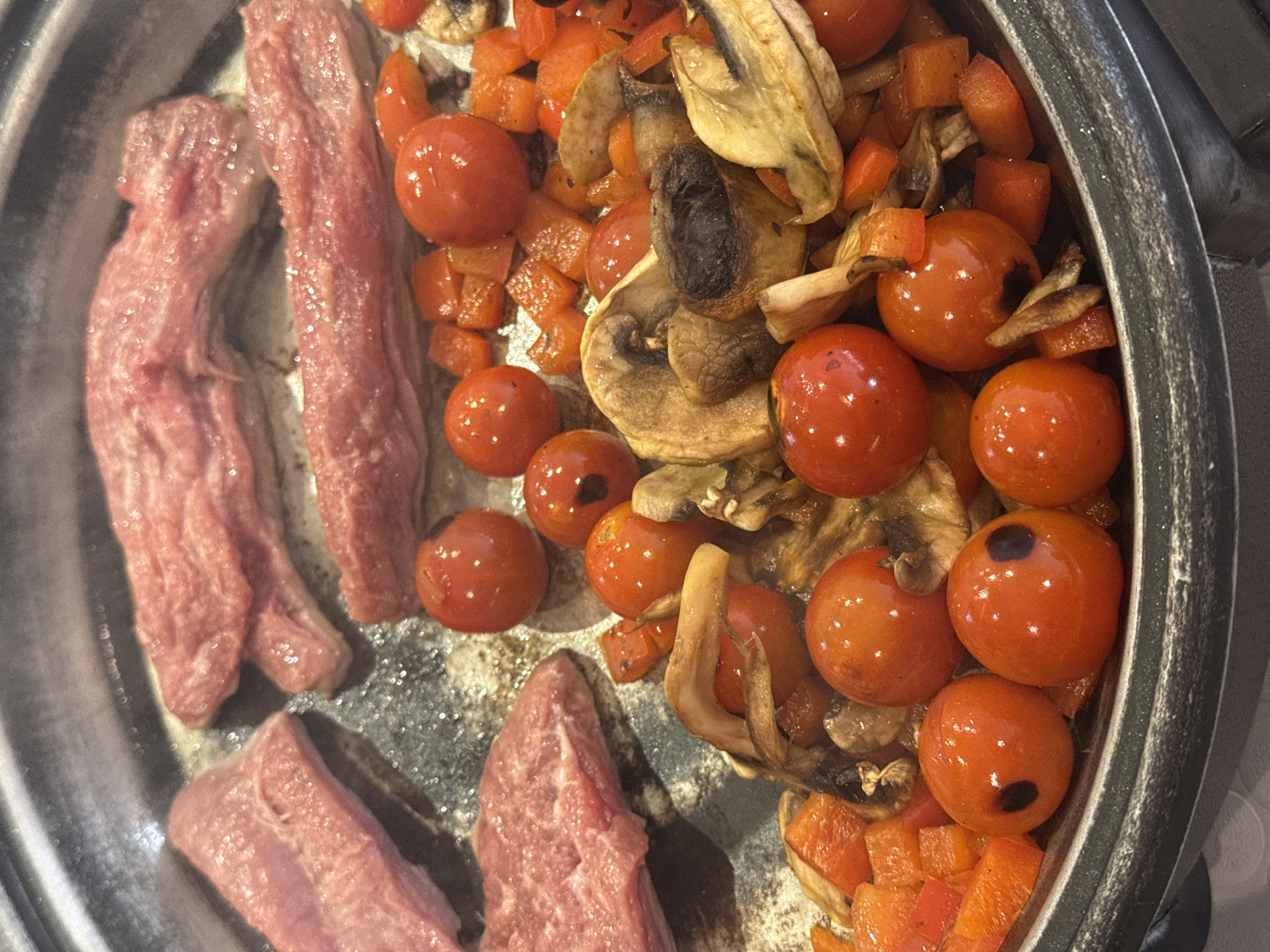 The image shows a cooking pan with various ingredients, presumably in the process of being prepared. There are pieces of raw meat, likely pork or beef, situated on one side of the pan. The other side of the pan contains a mixture of vegetables, including whole cherry tomatoes, diced red peppers, and sliced mushrooms. The vegetables appear to be sautéed or lightly browned. The pan itself appears to be well-used, with some discoloration or staining. The image does not provide any information about San Bernardino Tlaxcalancingo, Mexico or any people, actions, interactions, or environment beyond the pan and its contents.