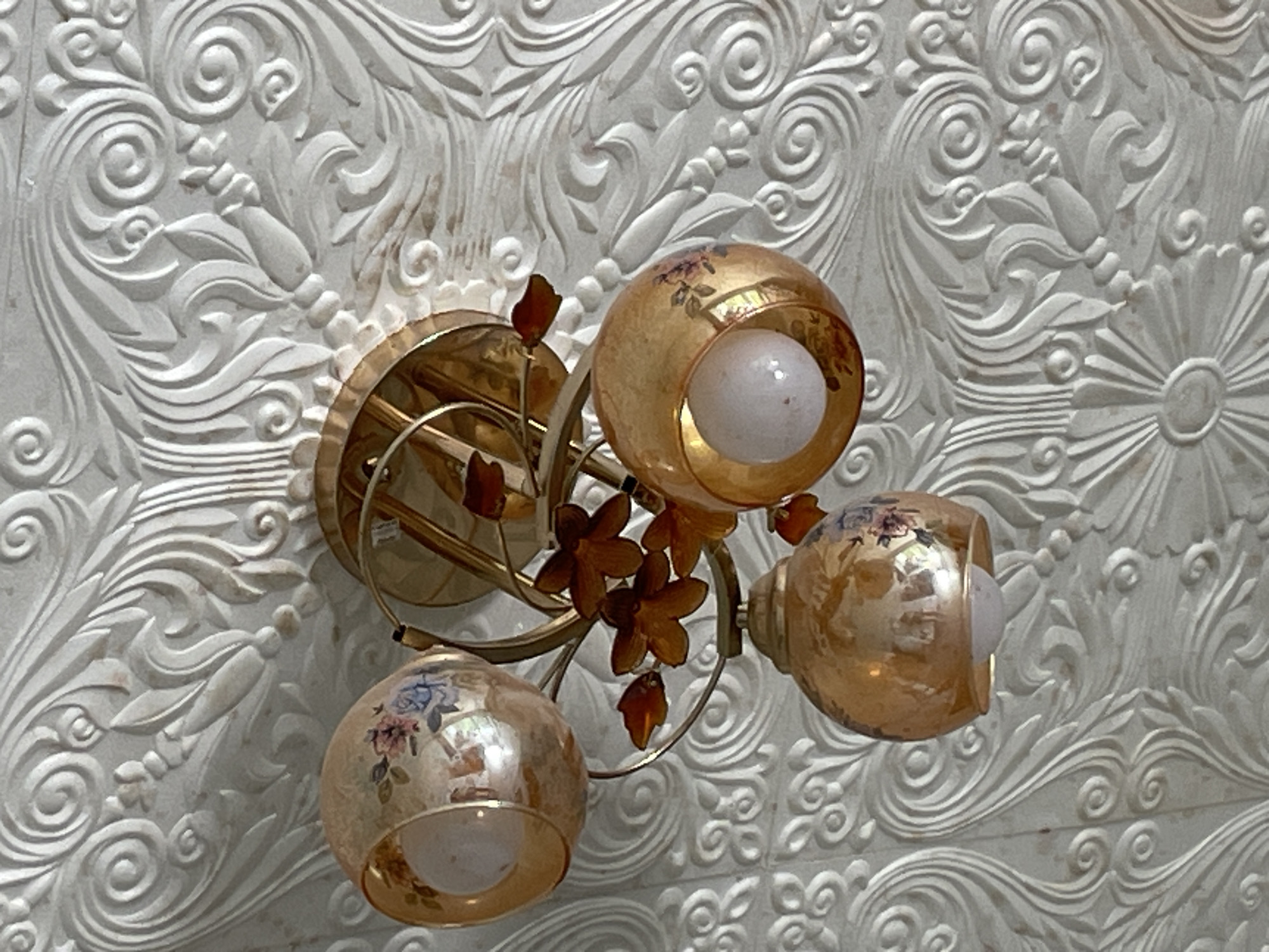 A three-light ceiling fixture with gold-toned shades decorated with floral motifs is mounted on a textured white wall. The fixture has a central base from which curved arms extend to hold each light shade. Small decorative floral accents are attached to the arms. The wall features a repeating pattern of stylized floral and swirl designs.