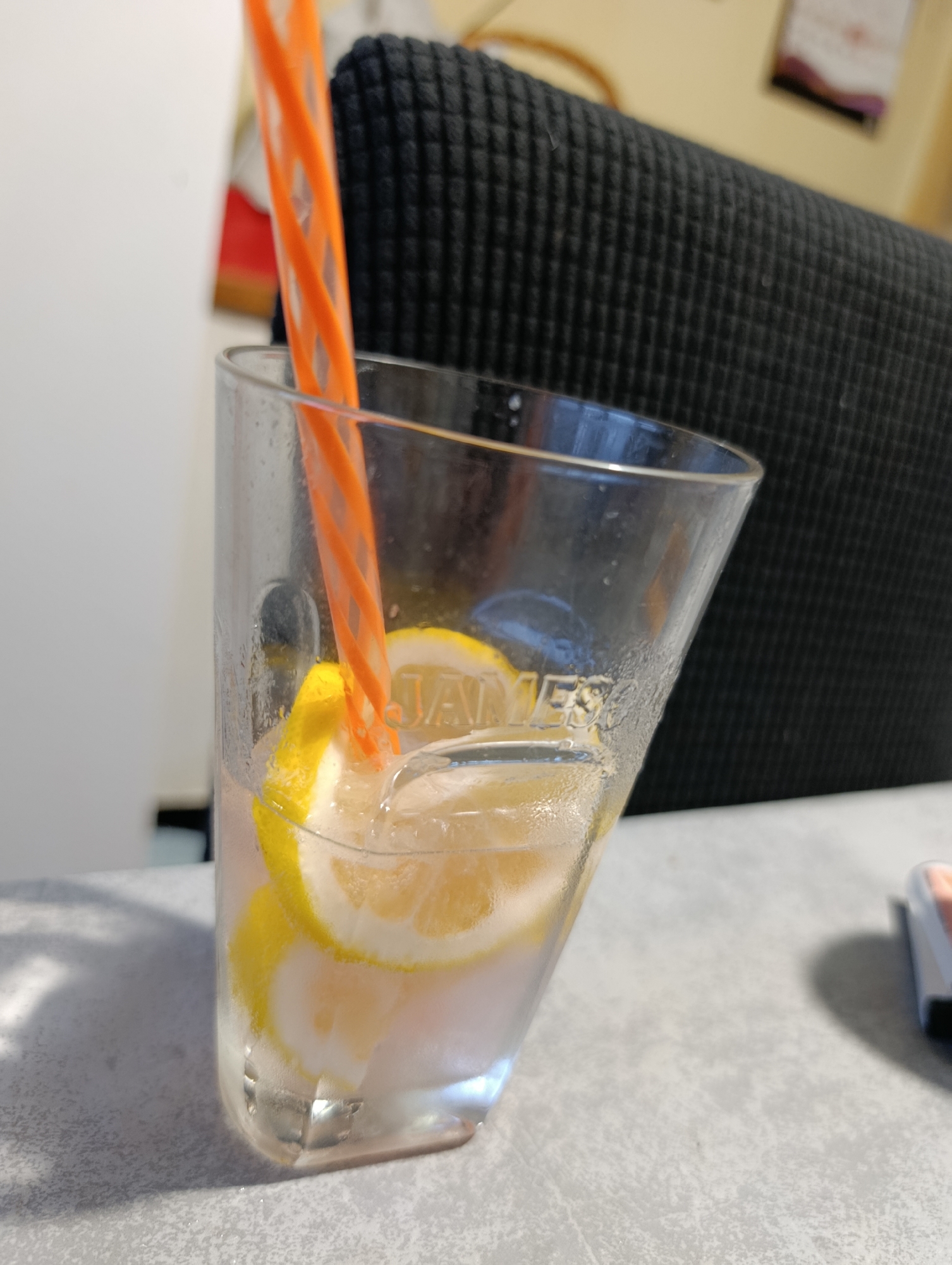The image shows a close-up of a glass of water with lemon slices and an orange straw. The glass is clear and has a slightly angled shape. There are several thin slices of lemon floating in the water, some of which are partially submerged. The orange straw is coiled and has a spiral pattern, inserted into the glass from the top. The glass is sweating, indicating it contains a cold beverage.
In the background, a dark, textured surface, likely a chair back, is visible. To the left of the glass, a white wall or surface is out of focus. To the right, a portion of a gray surface, possibly a table, is visible, with a blurred object that appears to be a small electronic device or case.
The lighting suggests an indoor setting, possibly during the daytime, with soft light illuminating the scene. The overall impression is that of a refreshing drink prepared for consumption. There is some visible text on the glass, but it is partially obscured and difficult to read. It appears to be a brand name or logo.