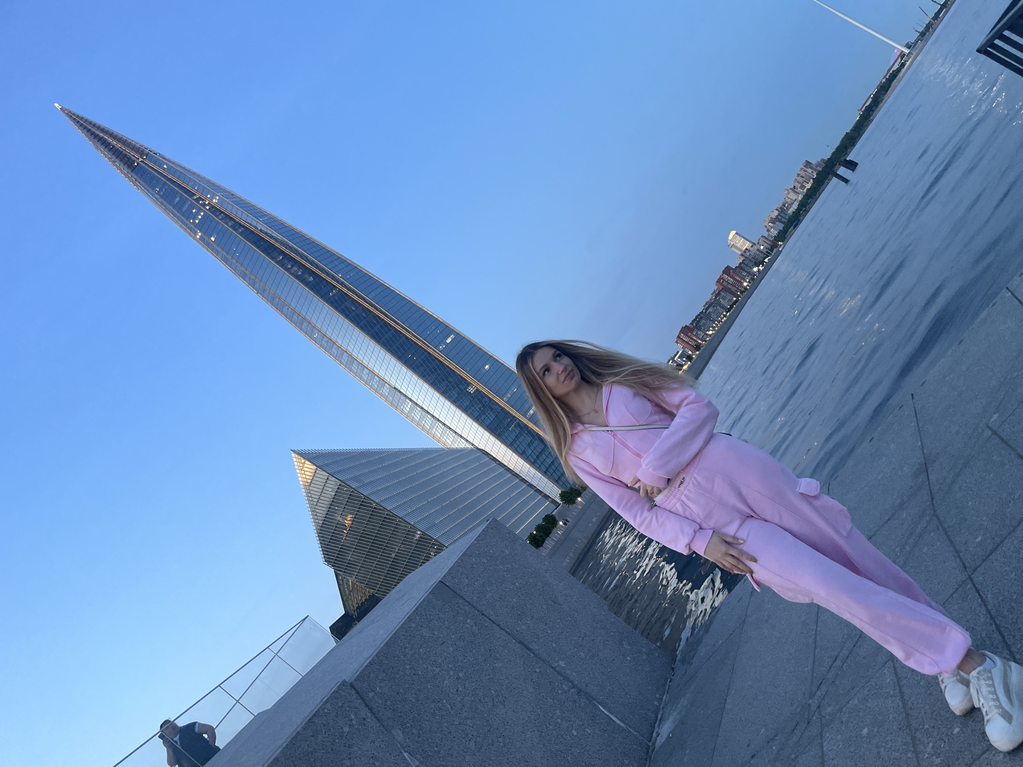 A young woman stands on a paved walkway near a body of water, with a distinctively shaped skyscraper dominating the background. The woman is wearing a pink tracksuit and white sneakers. She is looking to her left, with her hand on her chest. The skyscraper, the Lakhta Center, is a prominent landmark in Saint Petersburg, Russia, and its distinctive needle-like structure is visible against a clear blue sky. The walkway appears to be part of a waterfront promenade, with a stone wall to the left and the water stretching out to the right. In the distance, other buildings and a city skyline can be seen. The lighting suggests it is either early morning or late evening.