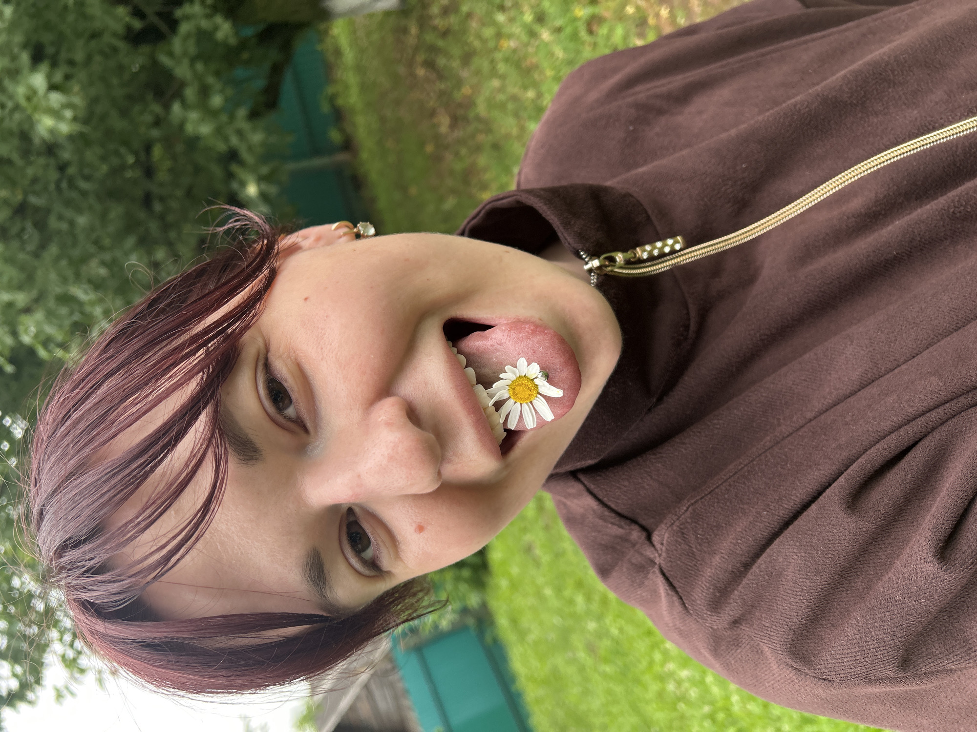 A person with dark red hair is lying down outdoors, with their face turned towards the camera. They are wearing a brown hooded sweatshirt. A small daisy is placed on their tongue. The person's eyes are open, and they are looking directly at the camera with a playful expression. The background consists of green grass and trees, suggesting an outdoor setting, likely during the daytime. There are no visible text elements in the image. The location context provided is Фоки, Russia.