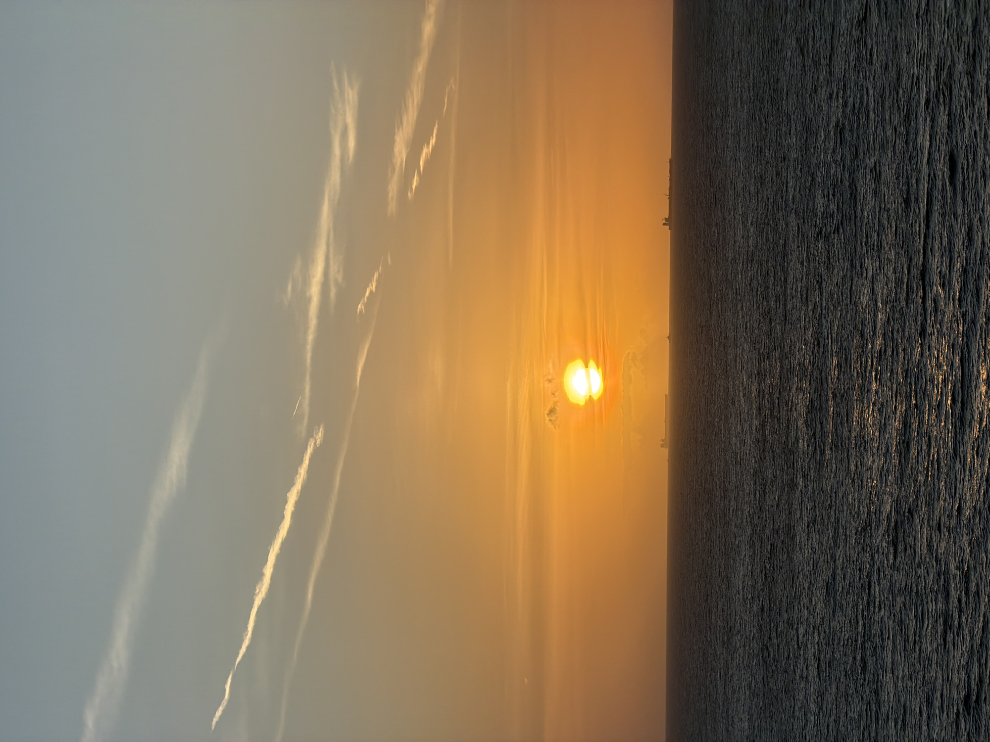 The image shows a sunset over the ocean. The sun is a bright orange orb, partially obscured by clouds near the horizon. The sky transitions from a light blue-gray on the left to an orange-yellow near the sun. Some wispy clouds and contrails are visible in the sky. The ocean surface is dark and textured, with some subtle reflections of the sunlight. A dark, barely visible coastline or cityscape appears on the horizon line. The image is rotated 90 degrees counterclockwise. The location is near a coastline in an unknown city and country.