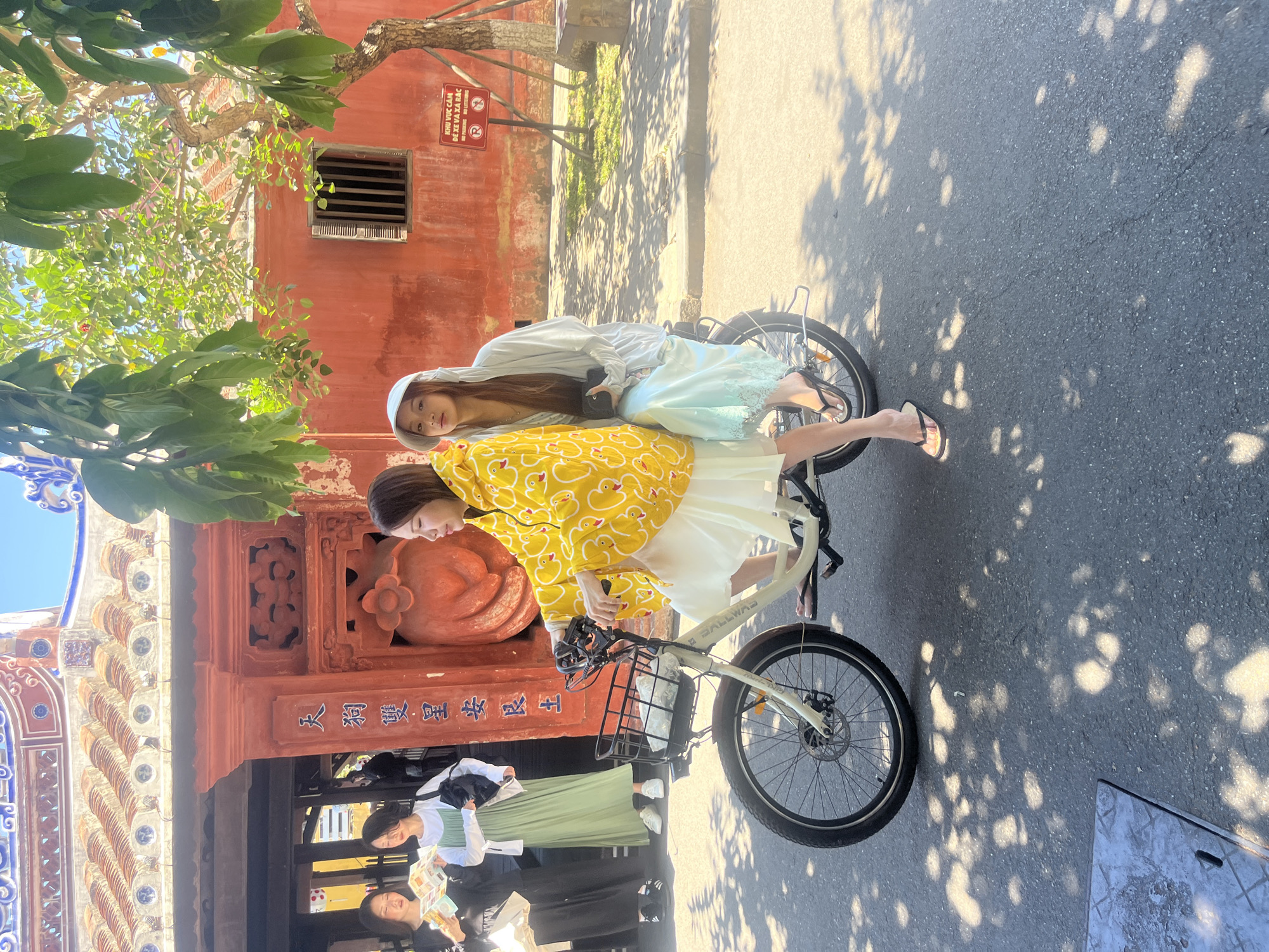 This image captures two young women posing on a white bicycle on a sunny day in what appears to be Hội An Ward, Vietnam.
The main subjects are two women. One, in the foreground, wears a vibrant yellow long-sleeved shirt adorned with white duck patterns and a white skirt. She is positioned on the bicycle, looking towards the left. Behind her, the second woman, wearing a light blue or mint green long skirt with a lace trim and a white hooded top, leans on her, also on the bicycle. Both women appear relaxed and are enjoying the moment. The bicycle is white, featuring a front basket.
The setting is an outdoor street scene with a distinct historical or traditional ambiance. In the background, there's a prominent reddish-brown building with intricate architectural details, including decorative carvings and a tiled roof structure. Further back, another red building is visible. Lush green trees and foliage are abundant, casting dappled shadows on the asphalt road, indicating bright sunshine. Other people can be seen in the background, some holding maps or brochures, suggesting a tourist-heavy area.
Visible text includes a red sign on the building in Vietnamese, which reads "KHU VỰC CẤM ĐỂ XE VÀ XẢ RÁC" (Area forbidden to park vehicles and litter). Chinese characters, "天狗雙星安艮土," are inscribed on the architectural structure of the foreground building, suggesting a temple or traditional hall. The brand "BALLWAS" is visible on the bicycle frame.
The image conveys a leisurely atmosphere, likely depicting tourists exploring the charming streets of Hội An under clear, sunny weather.