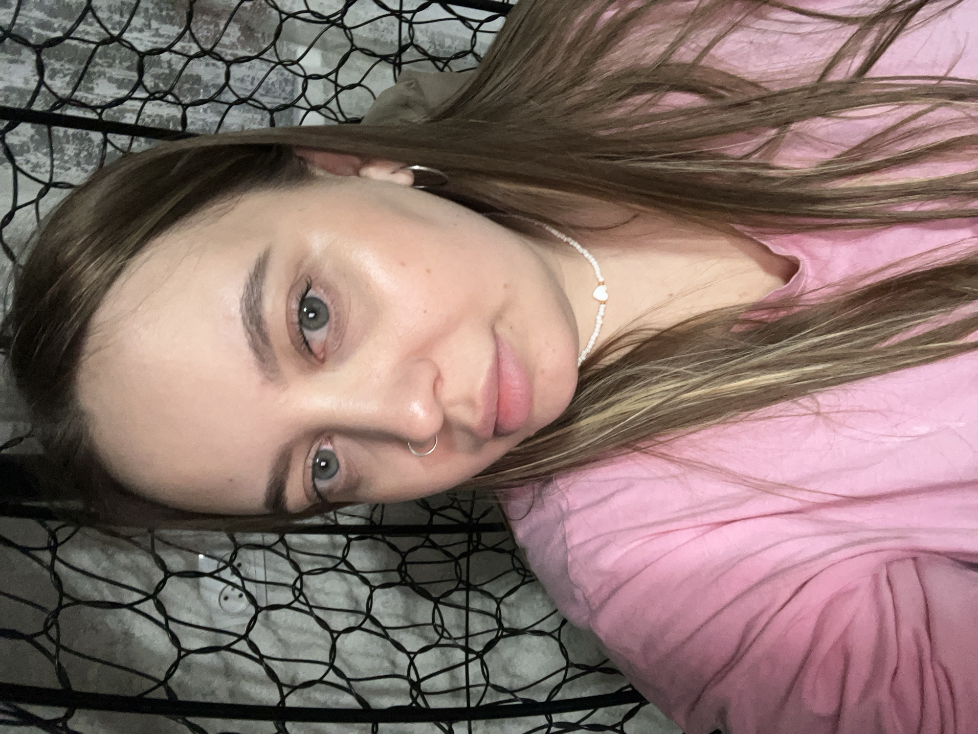 A woman with light skin and brown hair is lying down, possibly in a hammock or chair with a net-like structure. She is wearing a pink shirt and a beaded necklace with a heart charm. She has a nose ring and is looking at the camera. The background is a white wall. The location is not discernible from the image.