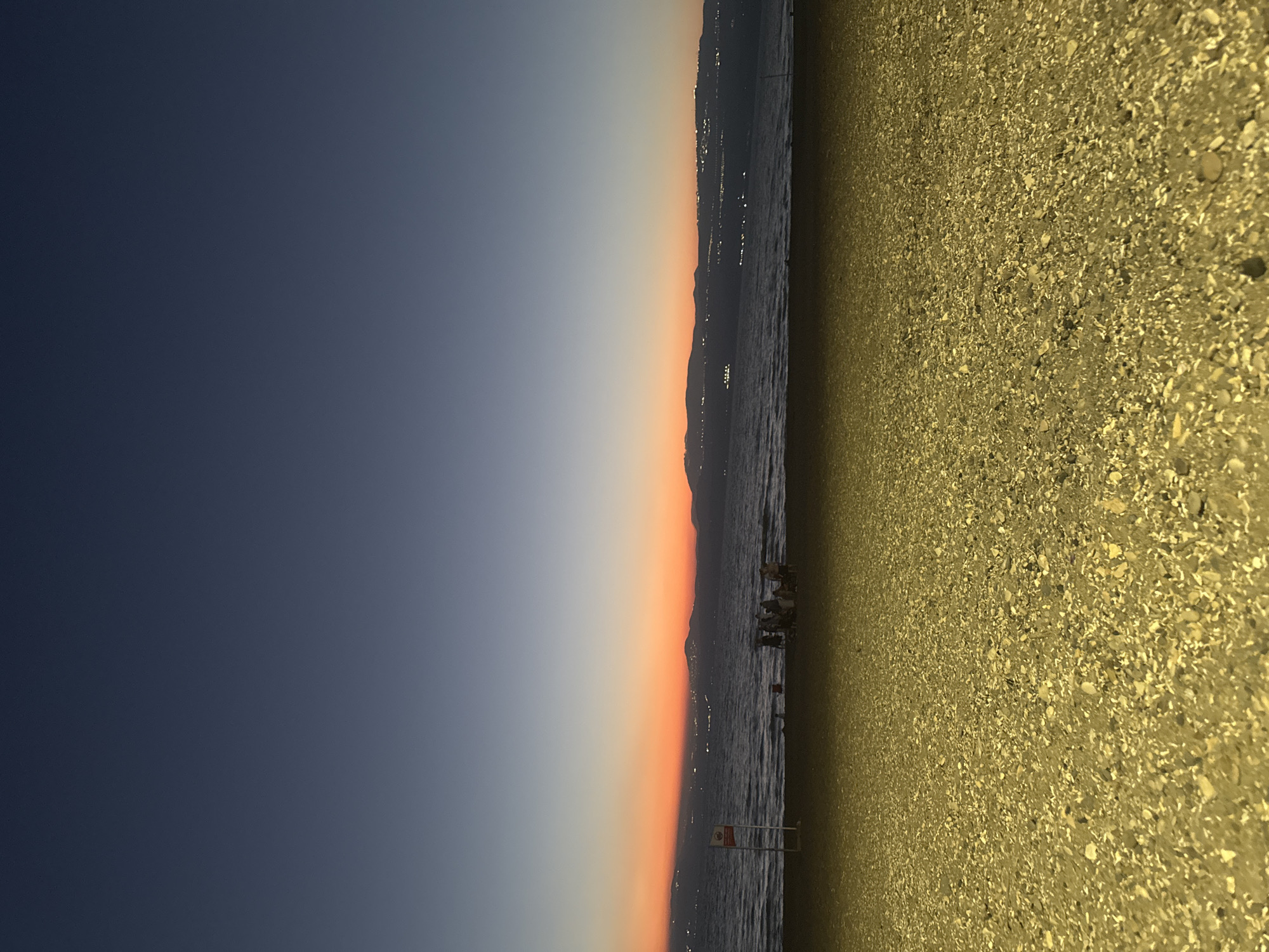 The image depicts a serene beach scene at dusk on the shores of the Sea of Galilee, located in the Golan Regional Council, Israel. The sky transitions from a deep navy blue at the top to hues of orange and pink near the horizon, indicating the afterglow of sunset. The calm sea stretches across the middle ground, with gentle waves lapping the shore. In the distance, the silhouette of rolling hills and mountains forms the opposite coastline, dotted with lights from settlements. A group of people are gathered on the sand near the water's edge, seemingly enjoying the twilight. The foreground is dominated by a textured beach composed of small pebbles and shells, illuminated by an artificial light source casting a yellow hue. A red and white signpost is partially visible on the left side of the beach. The overall atmosphere is tranquil and picturesque.