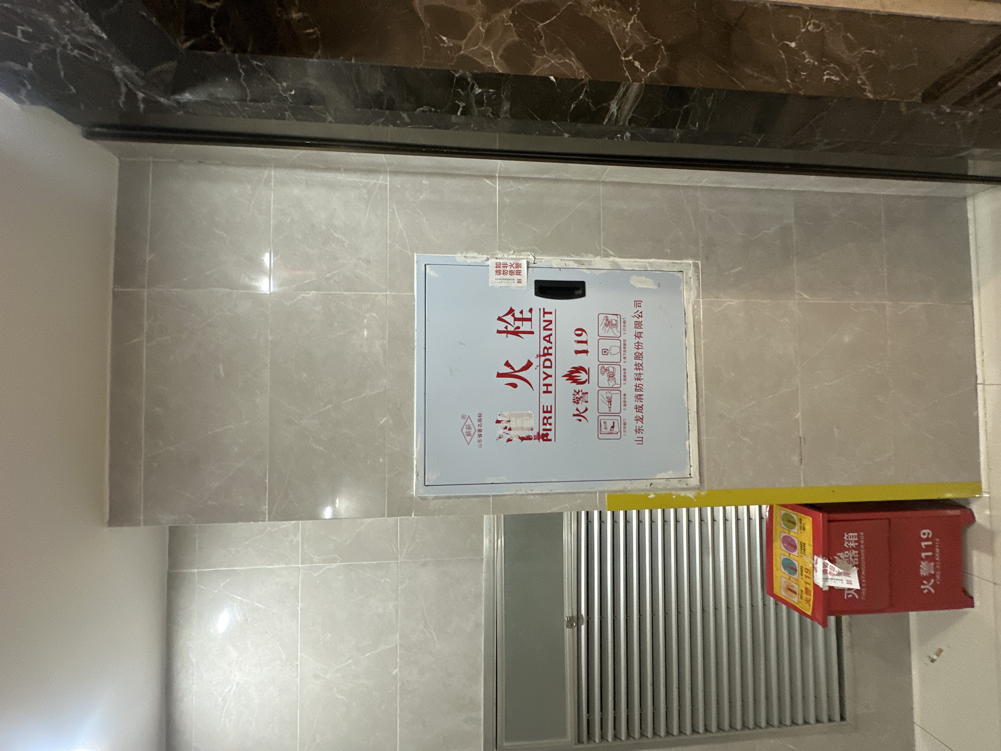 Here is a detailed description of the image:
The image shows a section of a wall in what appears to be an indoor space, possibly in the Chongqing High-tech Zone, China, given the Chinese text present. The wall is tiled with large, gray, marble-patterned tiles. A dark, perhaps black marble, trim runs along the top of the wall.
Centered on the wall is a fire hydrant cabinet, identified by Chinese and English text as "FIRE HYDRANT" and "消火栓". The cabinet is gray and bears pictograms and instructions, also in Chinese.
Below the fire hydrant cabinet and slightly to the right is a louvered metal door, likely for a utility space. A fire extinguisher box is located to the right of this door, labeled "FIRE EXTINGUISHER BOX" and "火警119" (Fire Alarm 119). It is red with instructions and pictograms.