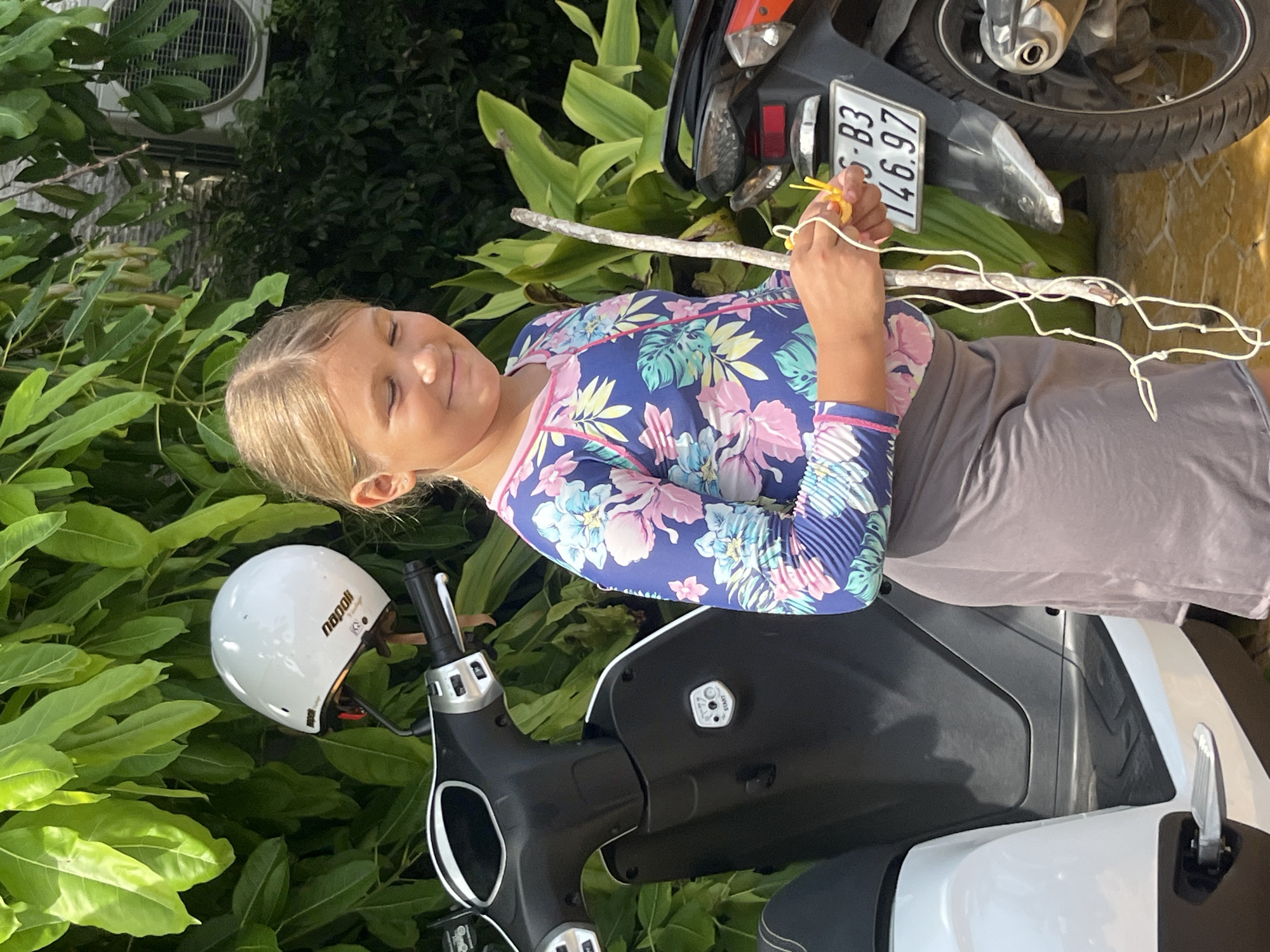 A young girl with blonde hair, wearing a blue and pink floral rash guard and a gray skirt, stands outdoors, holding a wooden stick with yellow and white string attached. She is smiling with her eyes closed and is positioned between a white scooter on her left and a black motorcycle on her right. A white helmet with "napoli" printed on it is hanging from the handlebars of the scooter. The background consists of lush green foliage and a section of an outdoor air conditioning unit. The ground is paved with yellow tiles. The license plate on the motorcycle reads "S-B3 146.97". The scene appears to be in the city of Phu Thuy Ward, Vietnam.