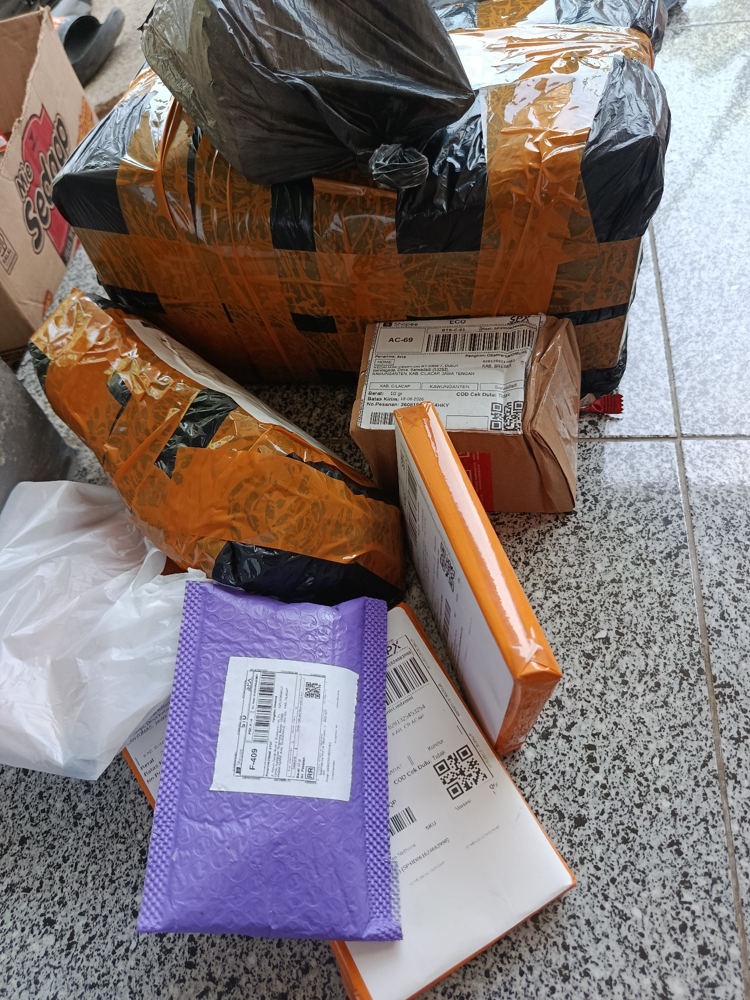 The image shows several packages on a tiled floor. The packages are wrapped in brown paper and orange tape, with some also covered in black plastic. One package has a label that reads "Shopee" and "ECO". Another label on a different package shows "AC-69" and "SPX". A small purple padded envelope is also visible. The background includes a cardboard box with the text "Mie Sedap" on it. The ground appears to be a grey tiled surface with a speckle pattern. There are no people visible in the image. The scene appears to be an outdoor or semi-outdoor area, possibly a porch or walkway, given the tiled flooring. The location is identified as Sarwadadi, Indonesia based on text visible on one of the shipping labels, which reads "KAB. CILACAP", "KAWUNGANTEN", and "DESA SARWADADI".