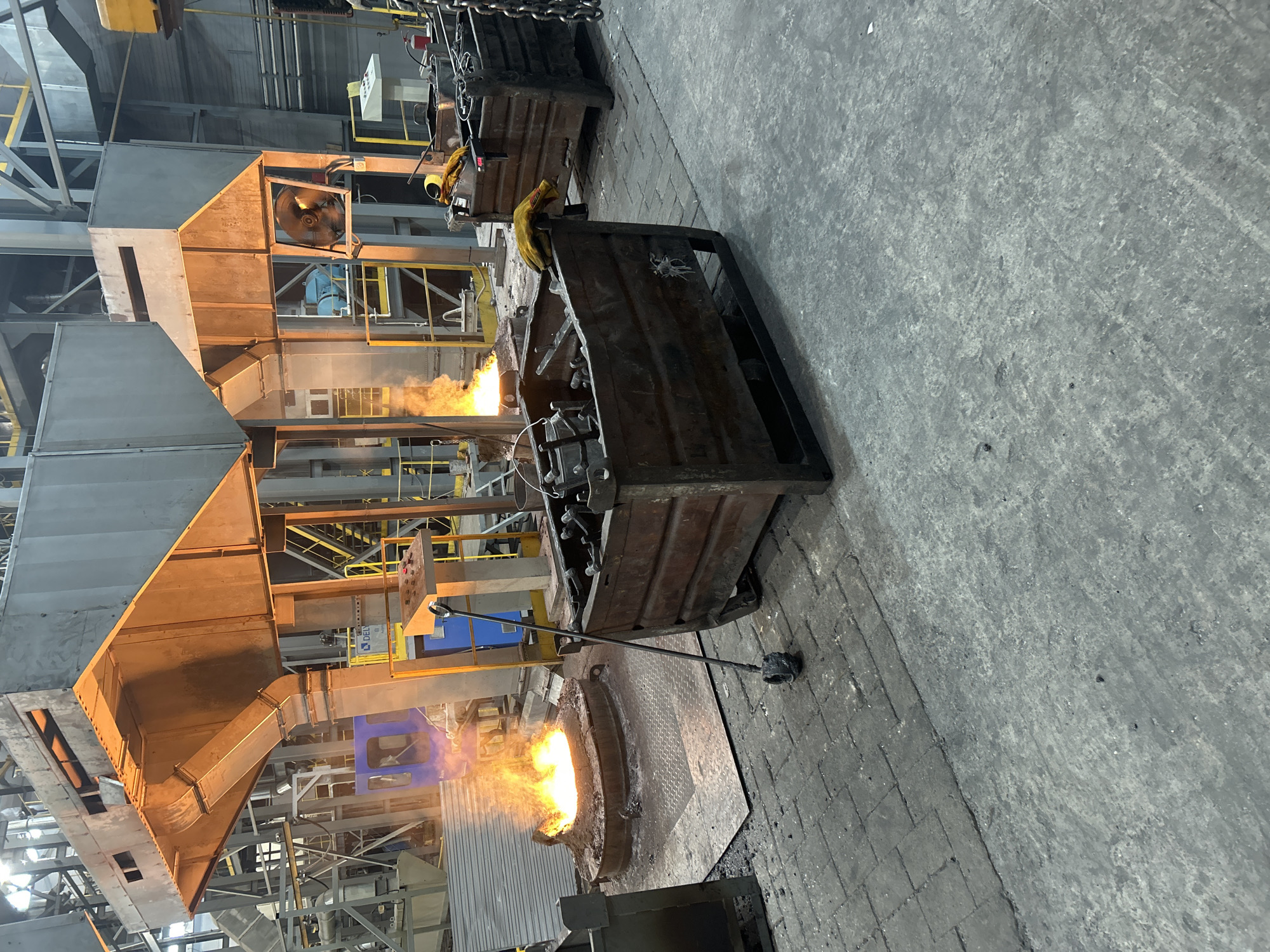 The image depicts an industrial setting, possibly a foundry or metalworking plant, with intense heat sources and heavy equipment. At the forefront, a circular furnace or crucible contains molten metal, evident from the bright orange flames emanating from within. A long-handled tool, presumably for manipulating the molten material, rests nearby. Metal containers filled with tooling sit behind the furnace. The floor is concrete or brick with a grimy, industrial patina. Overhead, a complex network of metal ductwork and structural supports dominate the upper portion of the image. The location is unknown and not related to the city of Noviy, Russia.