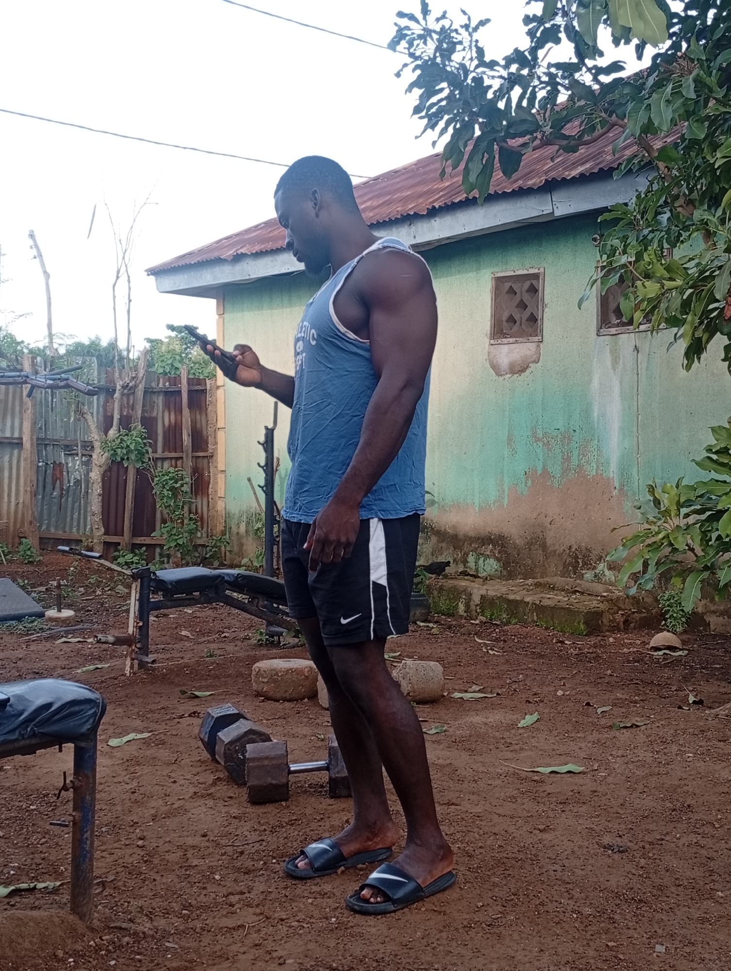 The image shows a dark-skinned man standing in an outdoor, informal gym setting in Akwanga, Nigeria. He is captured in profile, facing right, focused on a smartphone held in his right hand. The man has a muscular build, dressed in a blue tank top with partially visible white text that appears to say "ATHLETIC", black athletic shorts featuring a white stripe and a Nike swoosh logo on the left thigh, and dark slide sandals.
The ground is reddish-brown dirt. To the left, a black weight bench with a metal frame is visible, and further back, a pair of rusty, dark hexagonal dumbbells rests on the ground. A corrugated iron fence and some sparse vegetation are in the mid-ground. In the background, there's a weathered house with light green painted walls, showing considerable wear and discoloration, topped with a rusty brown corrugated iron roof. Two small windows with decorative grilles are visible on the house's wall. Lush green foliage from a tree hangs into the frame from the right. The sky is bright and clear, suggesting it is daytime, possibly late morning or afternoon. The man's expression is neutral as he interacts with his phone, appearing to be taking a break or preparing for exercise in this makeshift outdoor gym.