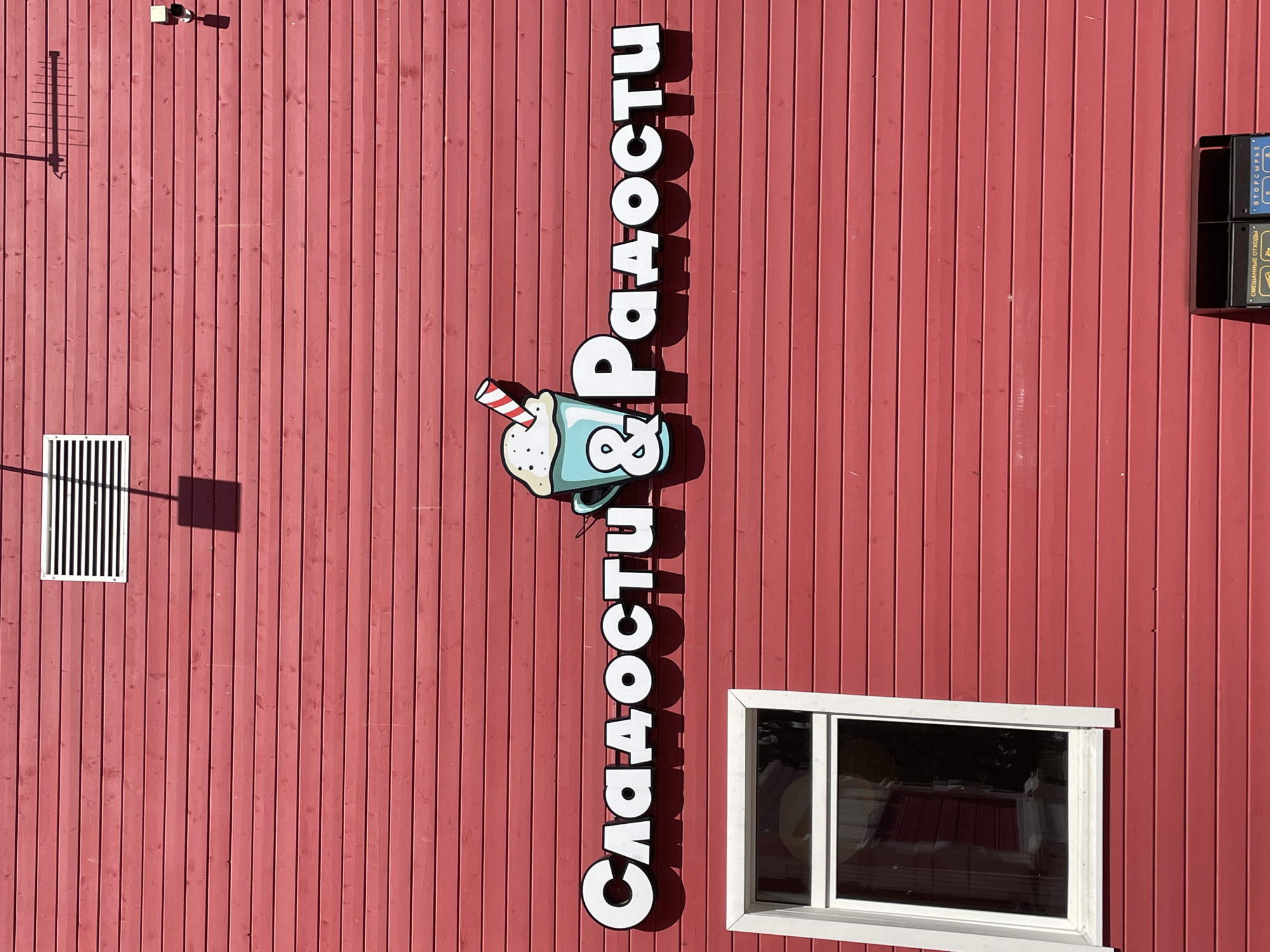 The image shows a close-up, eye-level, full shot of a bright red wooden building with a white-framed window on the left side. The building's exterior is constructed of horizontal wooden planks. A prominent sign with white, three-dimensional letters, reading "Сладости & Радости" (Sweets & Joys) in Cyrillic, is mounted on the building above the window. To the left of the "S" in "Сладости," there is a stylized cartoon image of a teal-colored cup with whipped cream and a red-and-white striped straw. To the right of the sign, an antenna and a security camera are visible on the red wooden wall. In the lower right corner, a portion of a black and yellow recycling bin system is visible, with labels for "СМЕШАННЫЕ ОТХОДЫ" (Mixed Waste) and "ВТОРСЫРЬЕ" (Recyclables). The lighting suggests it is daytime with bright sunlight, casting shadows from the sign and antenna onto the building's surface. The image was taken in the city of Vologda, Russia.