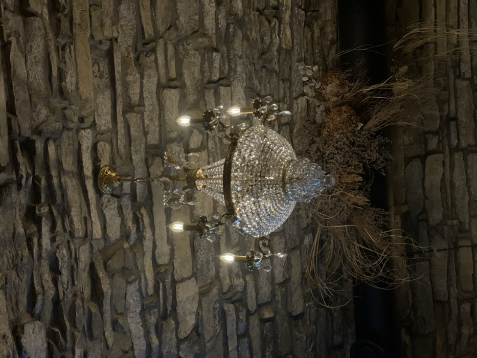 The image, rotated 90 degrees clockwise, displays an ornate chandelier hanging against a dark, textured wall. The chandelier is a prominent subject, featuring a dome or basket-like body entirely covered in numerous small, sparkling crystals that catch and reflect light. Several candle-style light bulbs are illuminated, casting a warm, inviting glow and highlighting the crystal elements. The chandelier's frame and suspension chain appear to be crafted from antique brass or a similar golden-toned metal.
The wall behind the chandelier exhibits a rough, uneven texture, resembling irregularly shaped stone blocks or concrete molded to mimic natural rock. Its dark gray and brownish hues provide a rustic, earthy contrast to the chandelier's elegant shimmer.
To the right of the chandelier, a large arrangement of dried botanical elements is visible. This includes various dried grasses, delicate branches, and other textured foliage, adding an organic and slightly wild aesthetic to the setting. A dark, possibly metal, pipe or structural column can be partially seen behind this dried arrangement.
The overall lighting is dim, suggesting an indoor evening setting where the chandelier serves as a primary light source, creating a cozy and atmospheric ambiance. No people are present, and no visible text can be discerned. The combination of the chandelier's antique elegance and the rustic stone-like wall, complemented by natural dried elements, evokes a historical or uniquely themed interior, fitting for a location in Kamianets-Podilskyi, Ukraine, known for its rich architectural heritage.