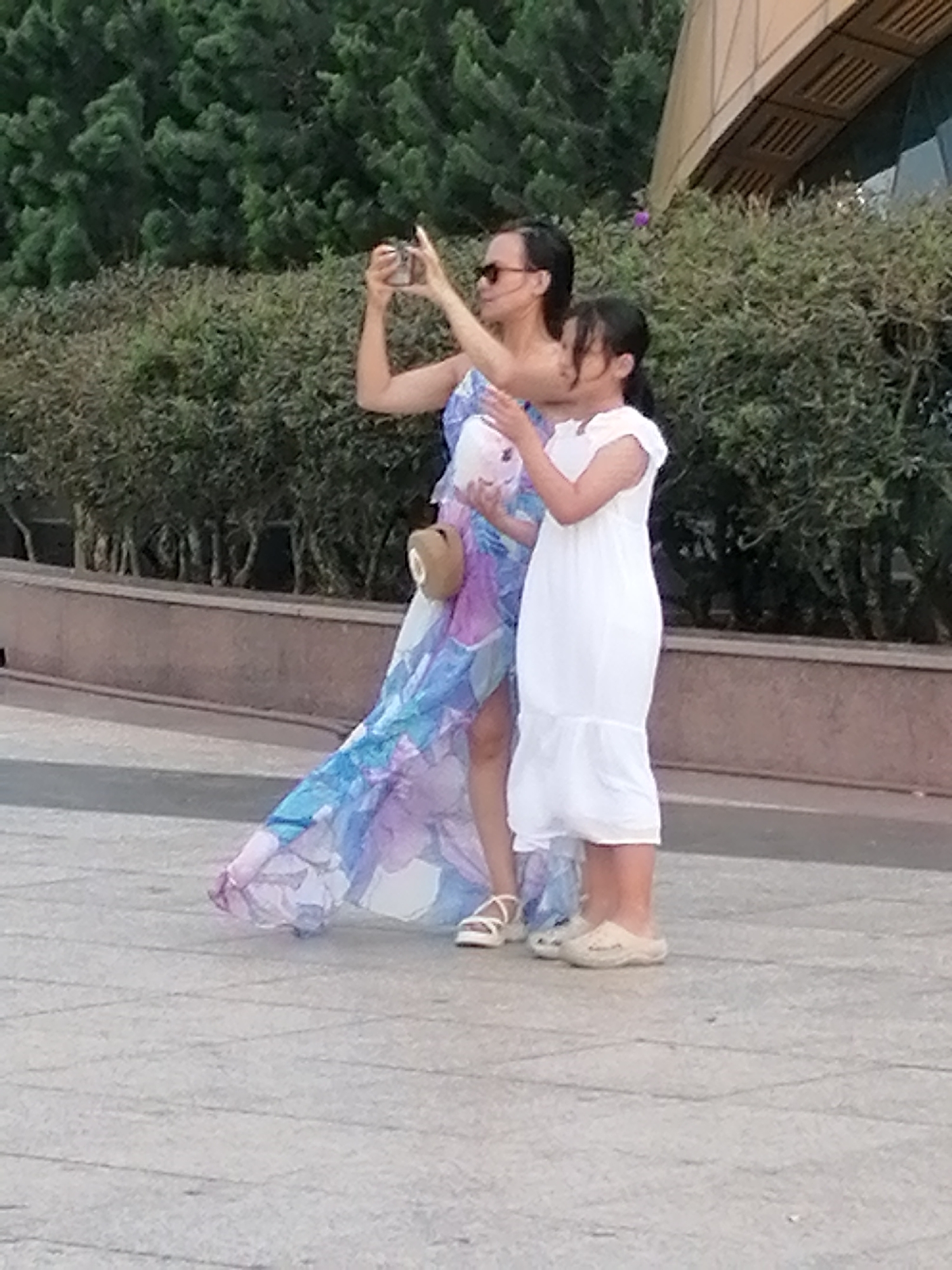 In Da Lat, Vietnam, a woman and a young girl are standing on a paved area. The woman, wearing sunglasses and a flowing blue and purple dress, is holding a phone up to take a picture. She has a light-colored bag slung over her shoulder. The girl, dressed in a white dress, stands beside her. Behind them, there is a line of green bushes and trees, and further back, a modern building with a curved, beige facade. The ground is paved with light gray square tiles.